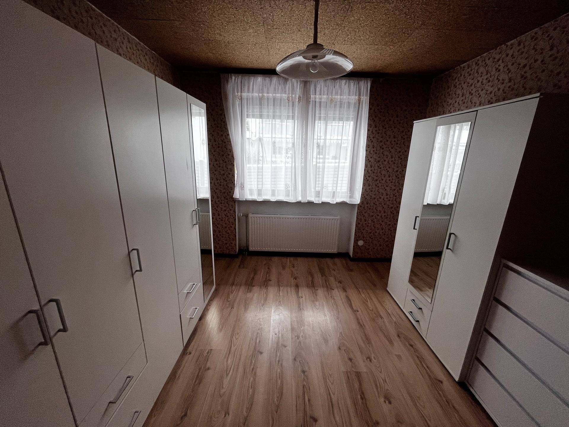 Schlafzimmer mit zwei Kleiderschränken, Holzboden und einem Fenster mit Gardinen.