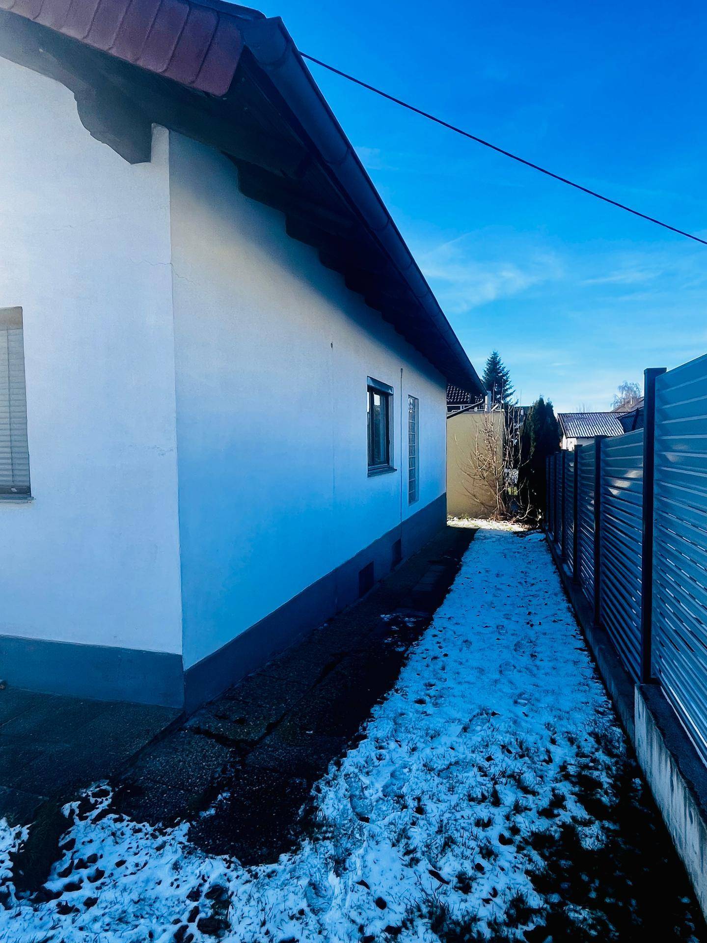 Schmaler Weg entlang der Hausseite mit Schnee und einem Zaun unter blauem Himmel.