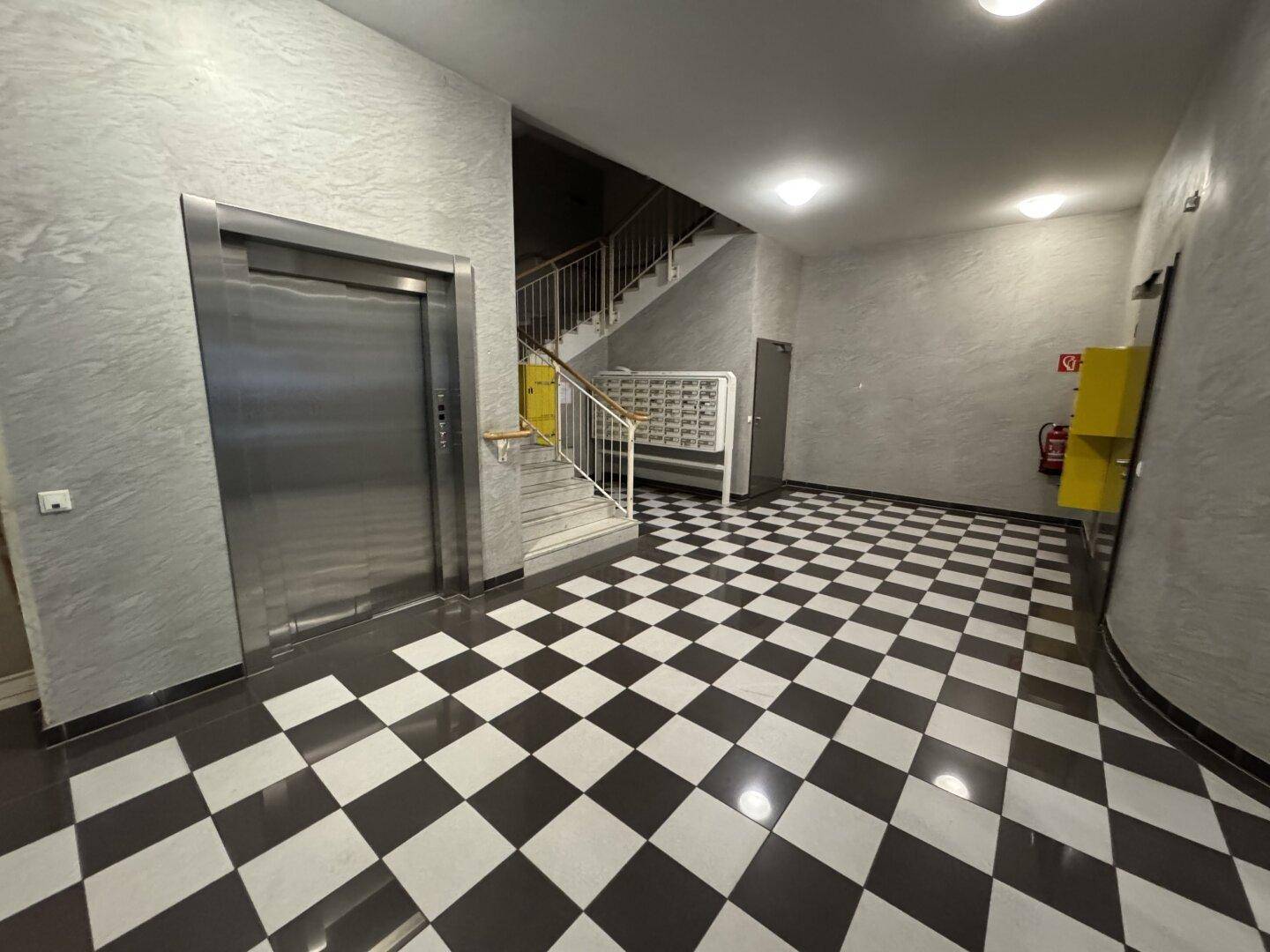 Eingangsbereich eines Gebäudes mit Aufzug, Treppe und schwarz-weiß kariertem Fliesenboden.