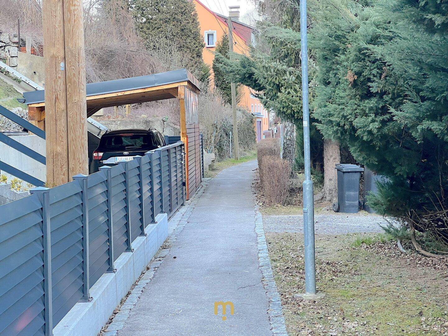 Schmaler Gehweg entlang eines modernen Zauns mit einem Carport und einem geparkten Auto im Hintergrund.