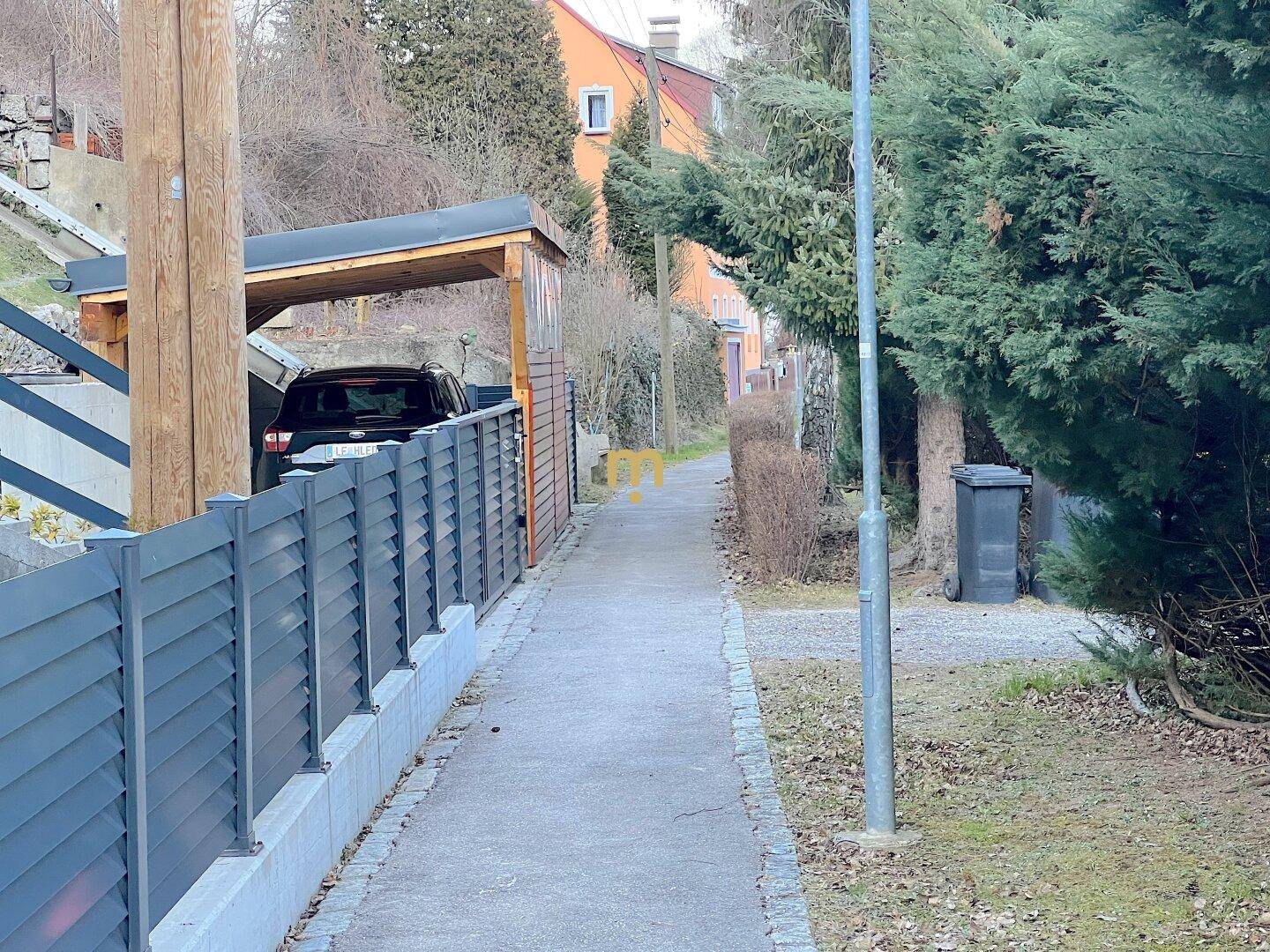 Gehweg neben einem modernen Zaun mit einem Carport und einem geparkten Auto im Hintergrund.