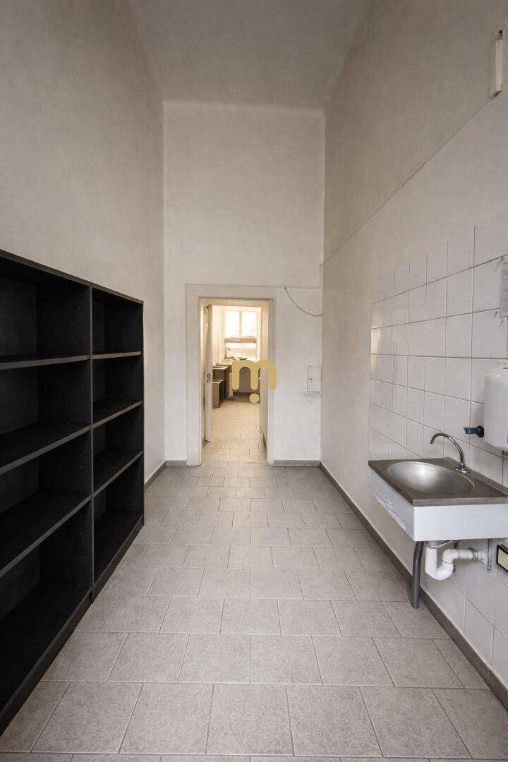 Ein schmaler Raum mit Regalen, einem Waschbecken und gefliesten Wänden, führt zu einem weiteren Raum.