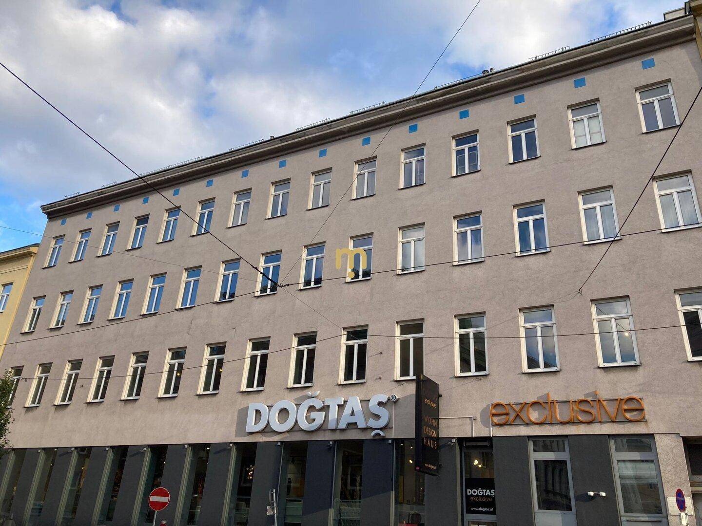 Gebäudefassade eines Mehrfamilienhauses mit vielen Fenstern und Geschäftsflächen im Erdgeschoss.