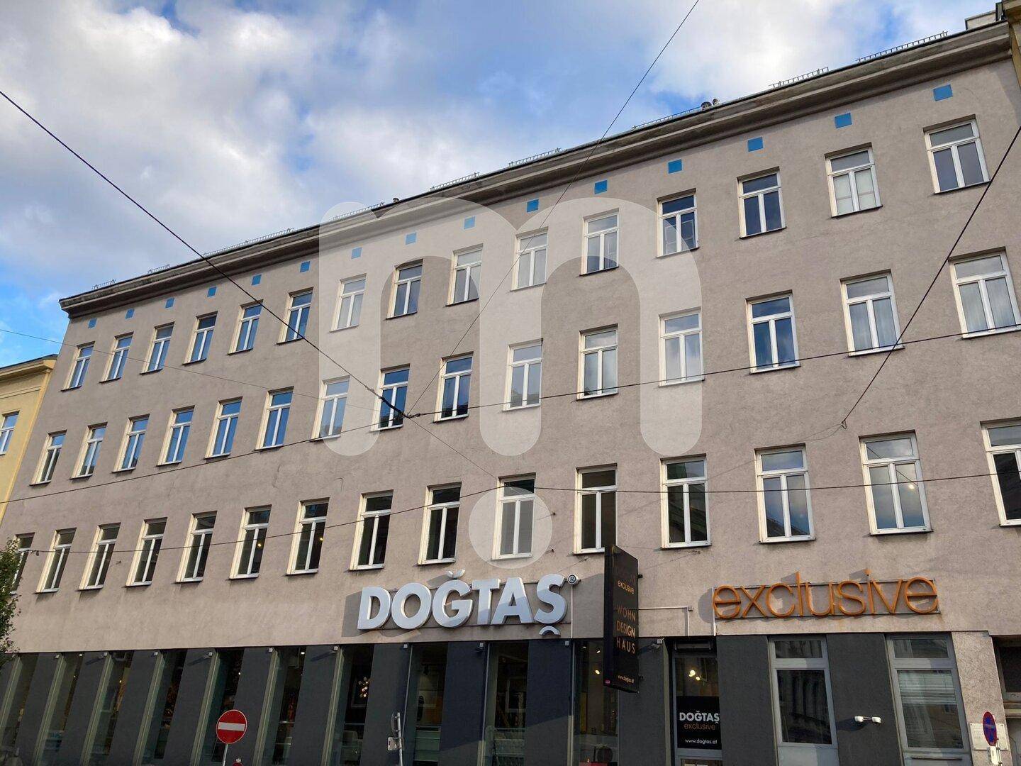 Mehrstöckiges Gebäude mit vielen Fenstern und Geschäftsflächen im Erdgeschoss unter bewölktem Himmel in der Stadt.