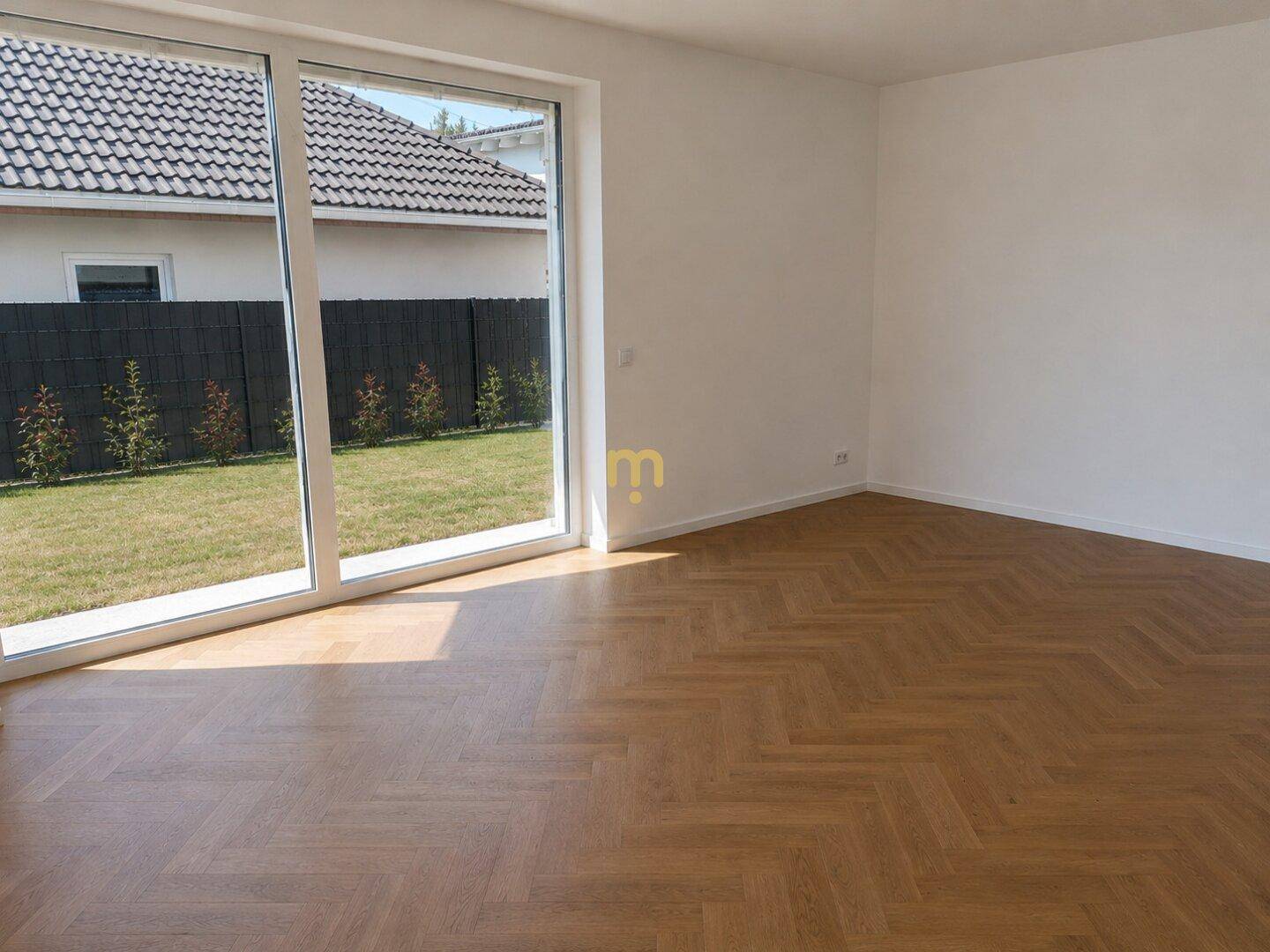 Heller, leerer Raum mit Fischgrätparkett und großen Fenstern zum privaten Gartenbereich.