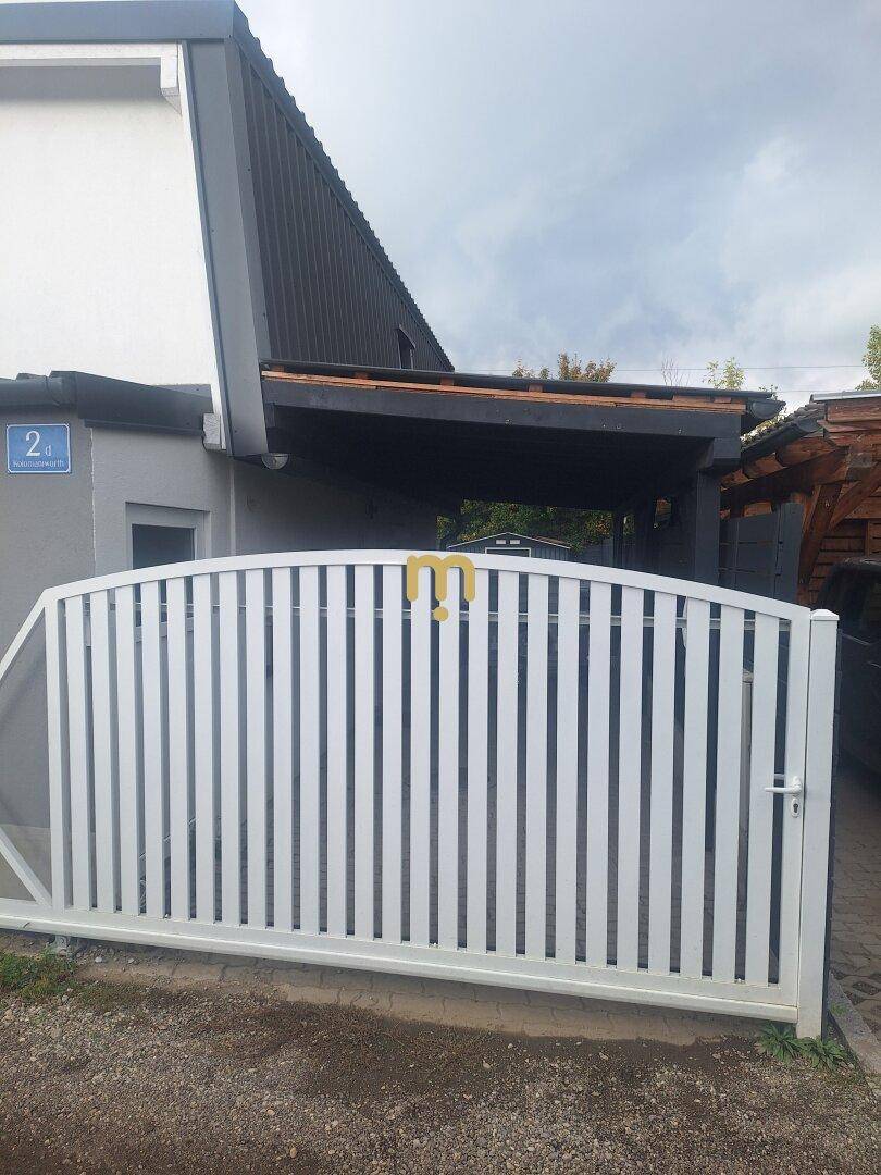 Modernes weißes Einfahrtstor vor dem Haus mit Carport im Hintergrund.