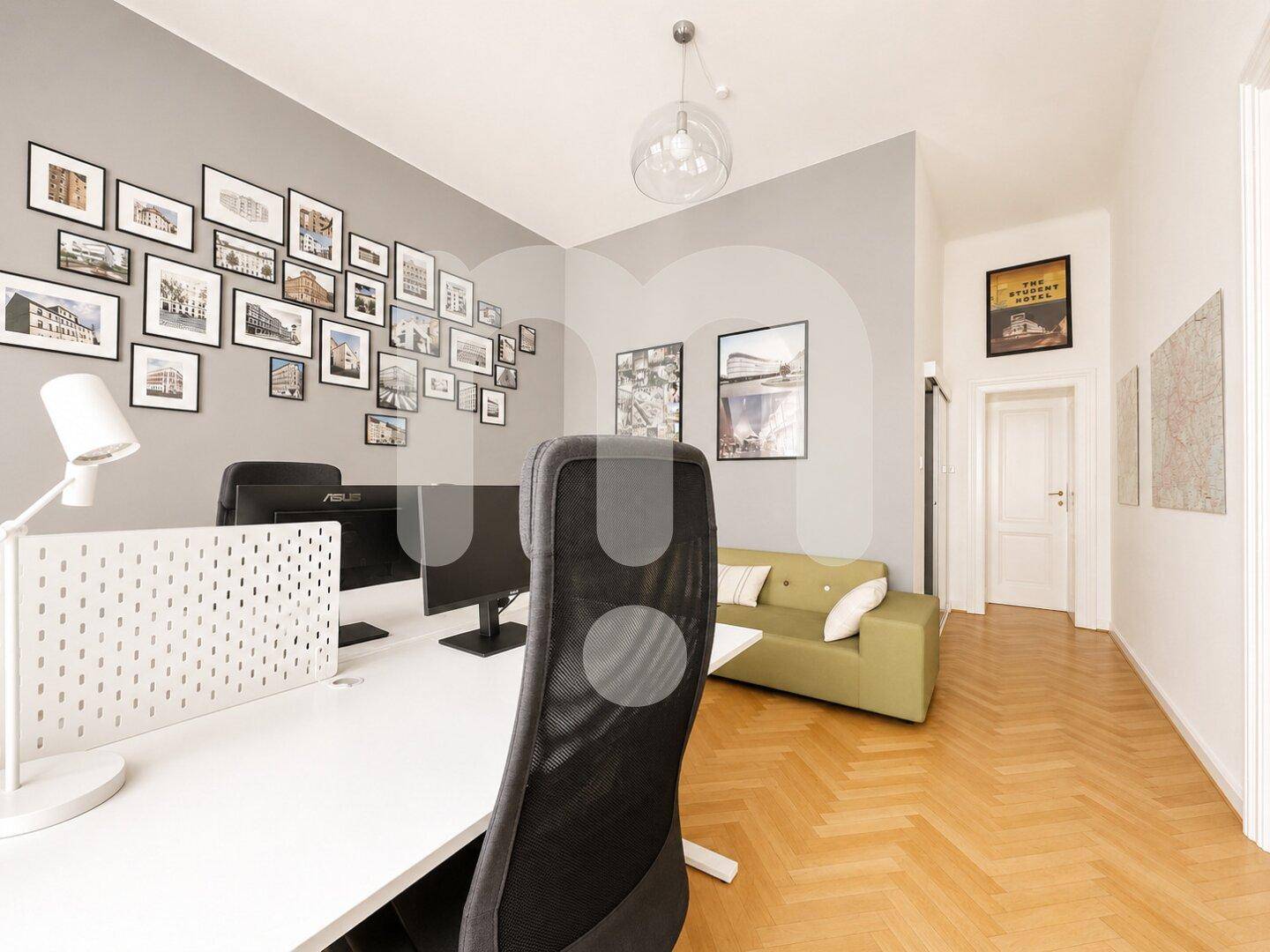 Arbeitsbereich mit Schreibtisch, Computer, grünem Sofa und einer Bilderwand, die zum Flur führt.