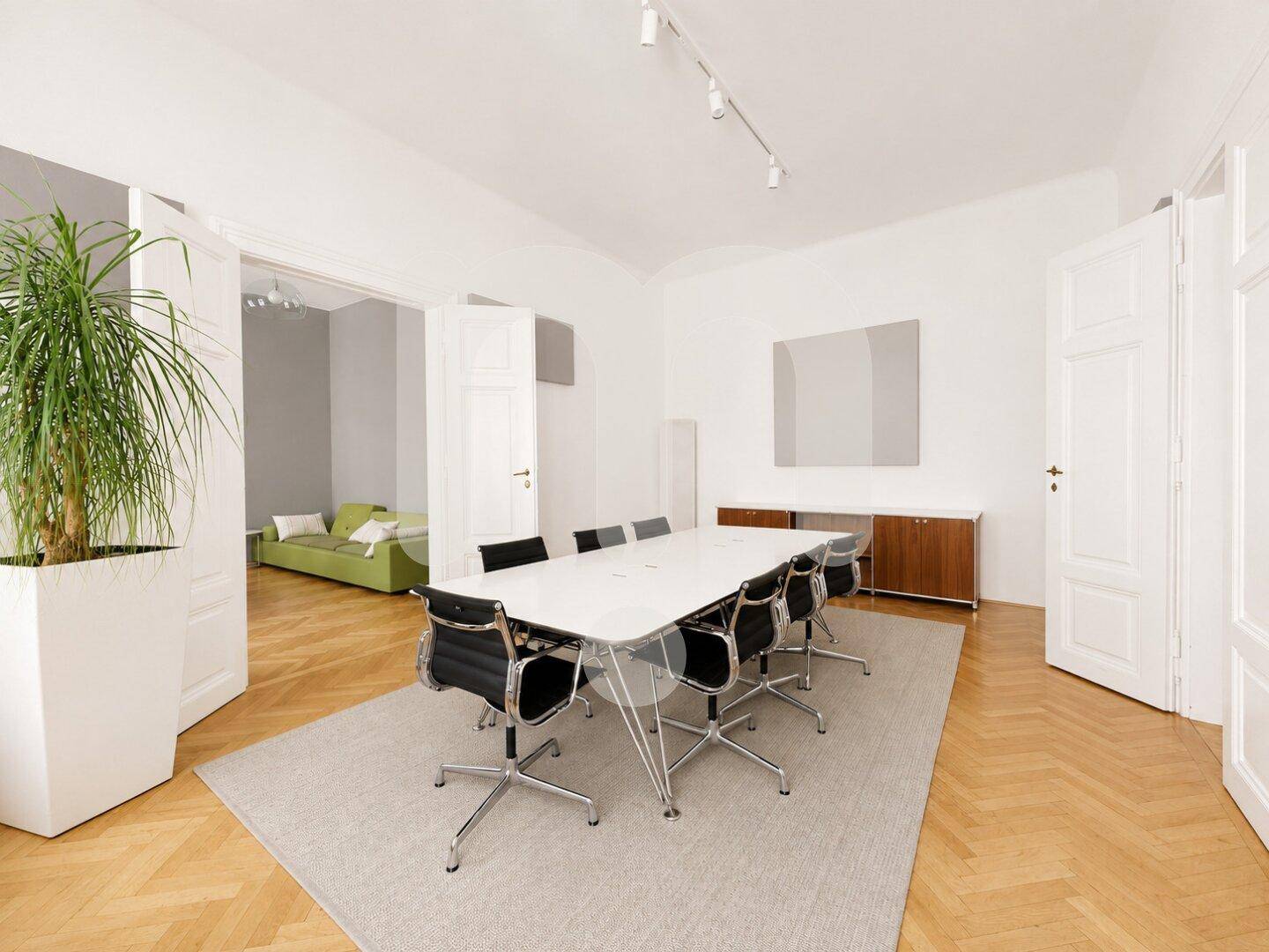 Heller Konferenzraum mit langem Tisch, schwarzen Stühlen und einem großen Pflanzenkübel, ideal für Besprechungen.