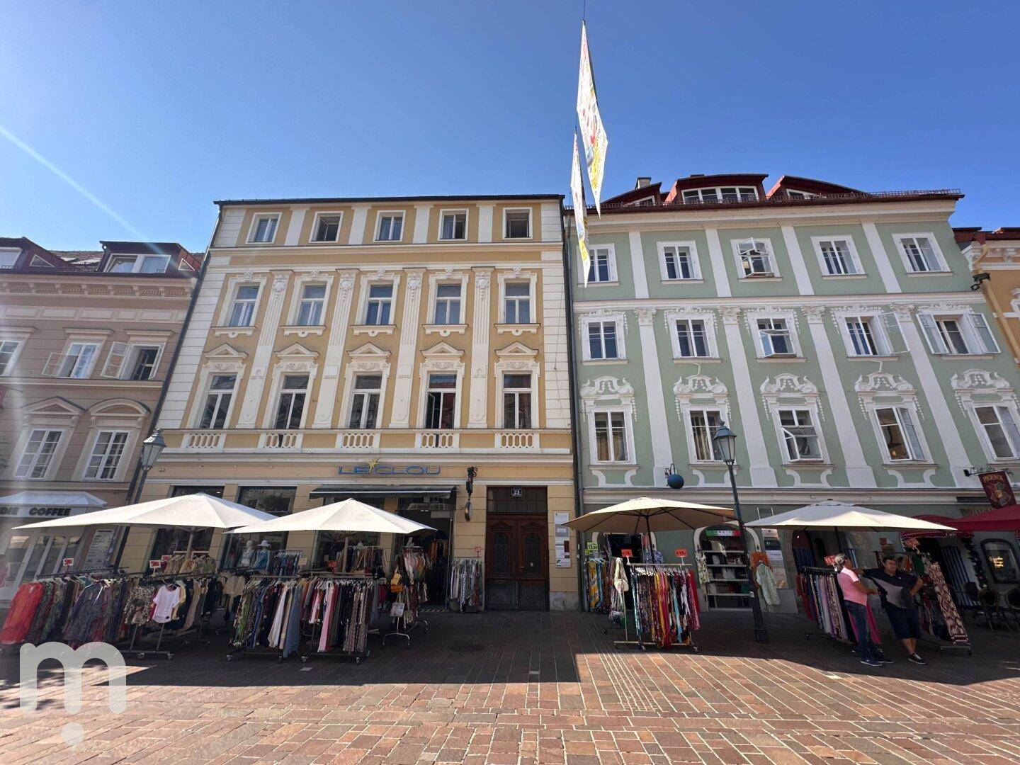 Historische Gebäude mit Geschäften und Marktständen auf einer belebten Straße unter blauem Himmel.