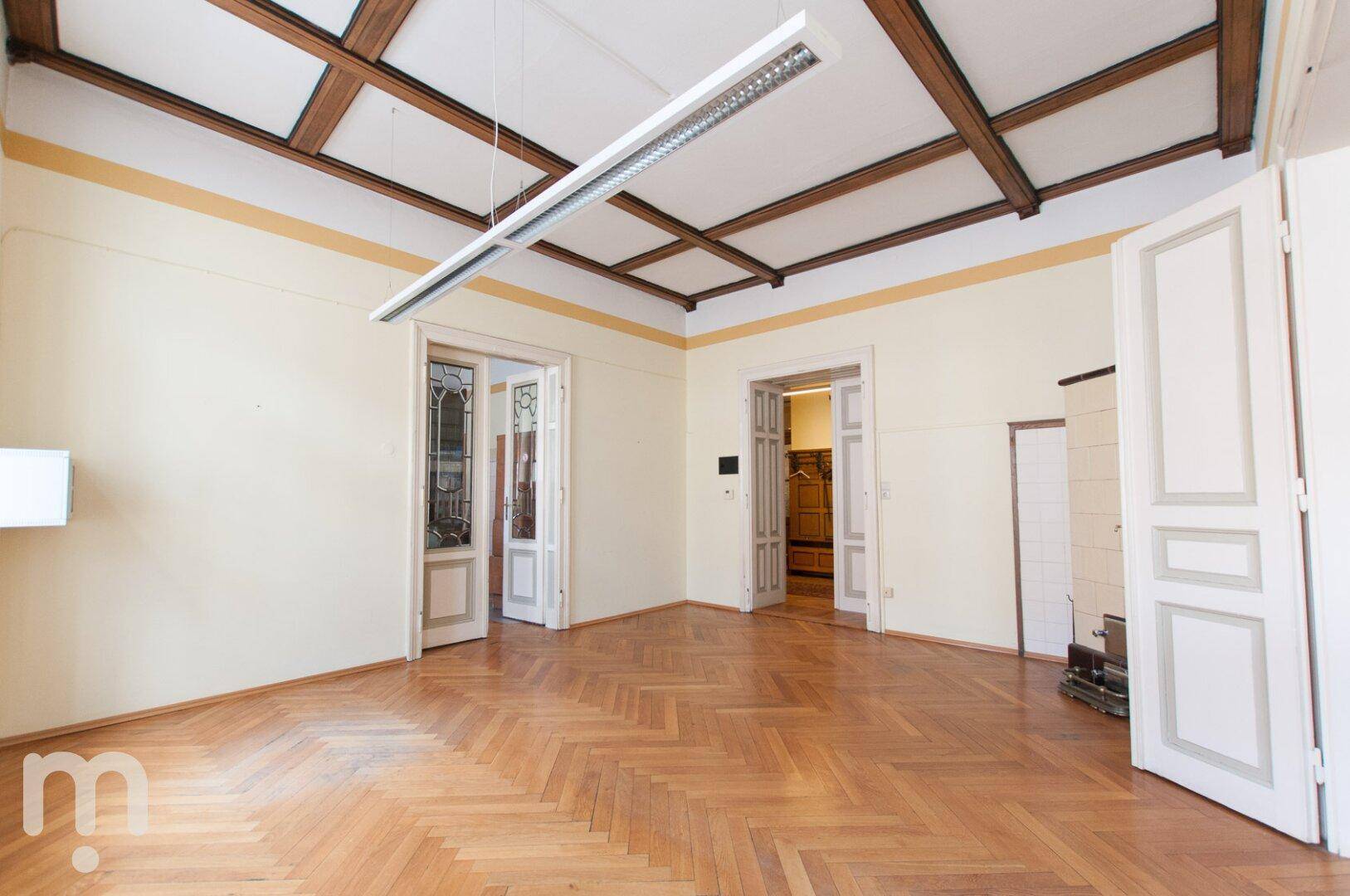 Ein geräumiger Raum mit Fischgrätparkett, hohen Decken und sichtbaren Holzbalken.