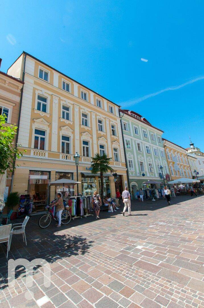 Eine sonnige Straße mit historischen Gebäuden, Geschäften und Passanten unter blauem Himmel.