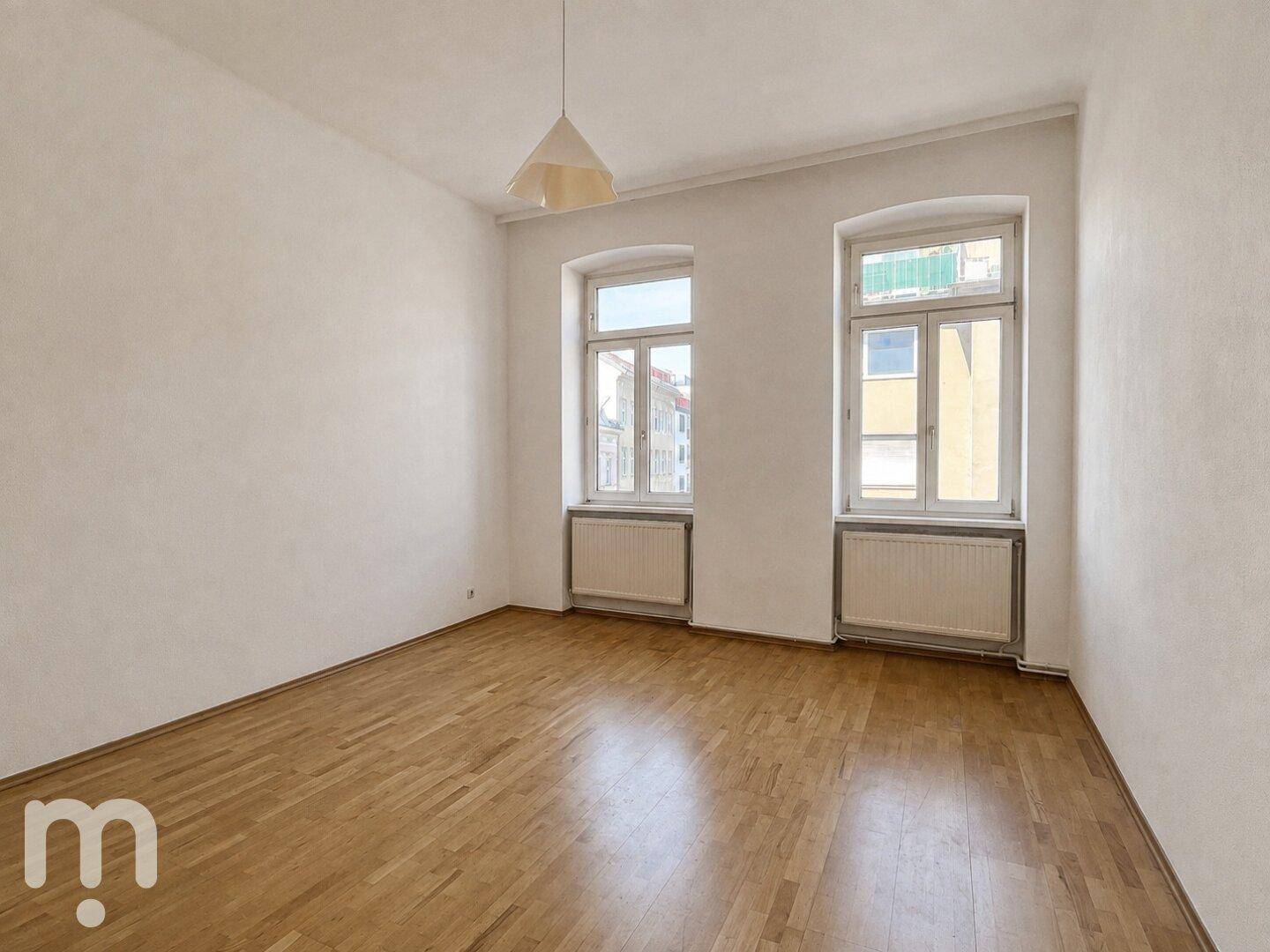 Leeres Zimmer mit hellem Parkettboden und zwei Fenstern, ideal für ein Schlaf- oder Wohnzimmer.