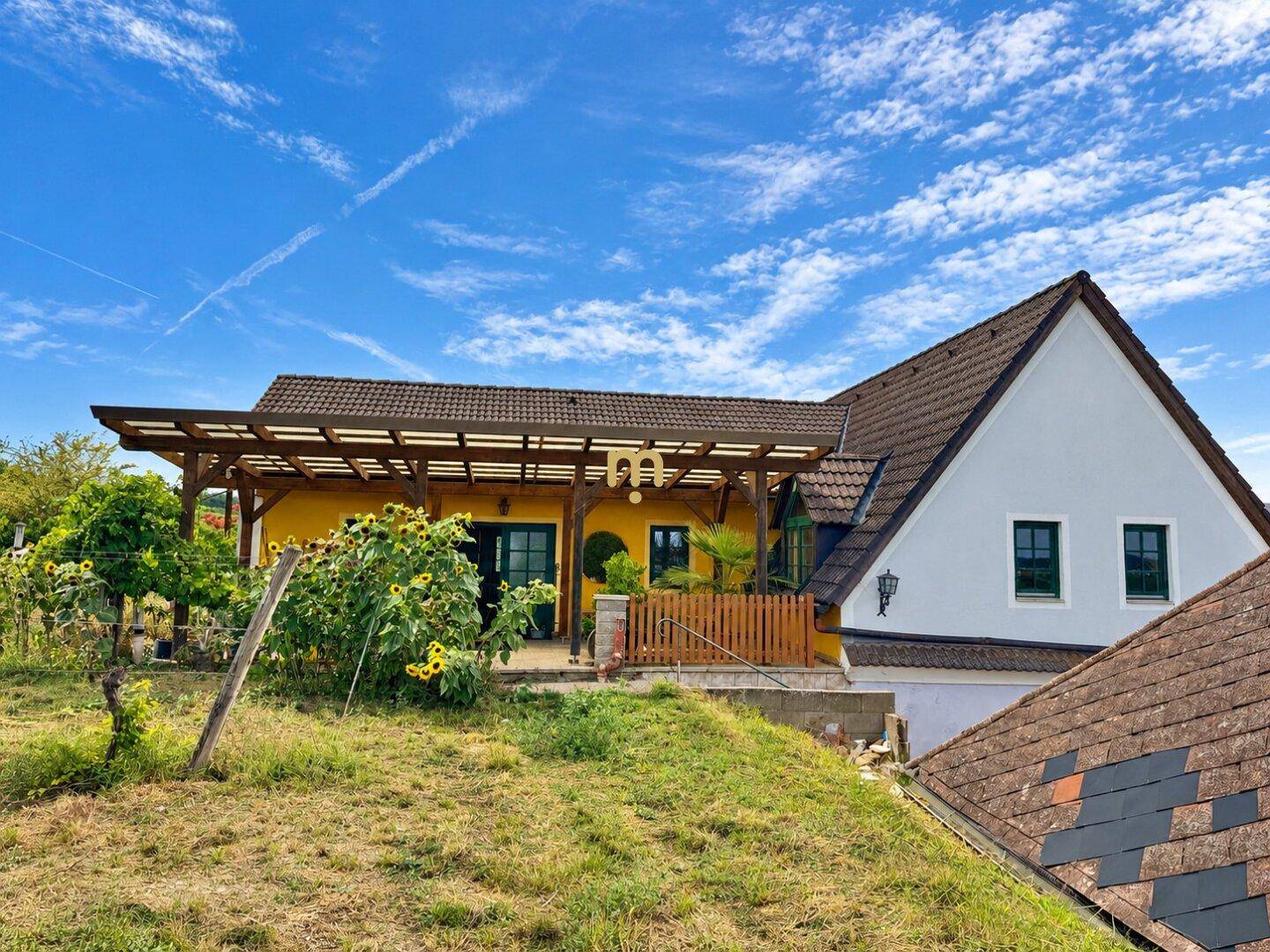 Ansicht des gelben Hauses mit überdachter Terrasse und dem angrenzenden grauen Gebäude unter blauem Himmel.