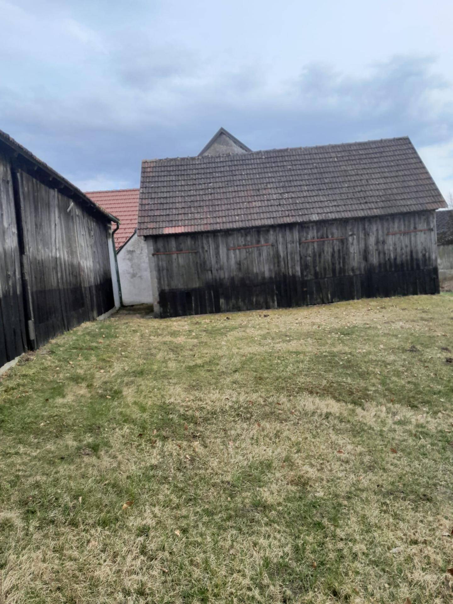 Zwei alte Holzscheunen mit Ziegeldächern und einem grasbewachsenen Bereich im Vordergrund unter bewölktem Himmel.