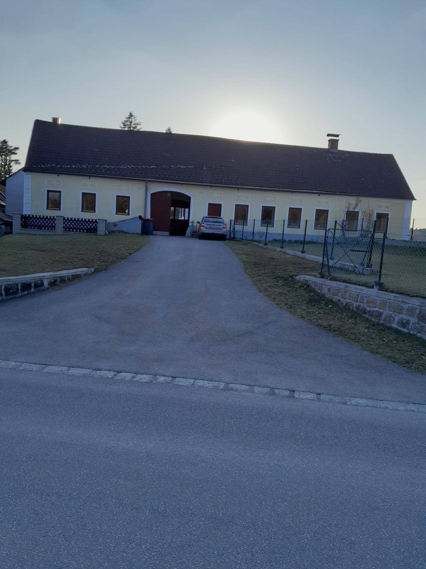 Vorderansicht eines gelben Bauernhauses mit großem Hof und Einfahrt, die Sonne scheint im Hintergrund.