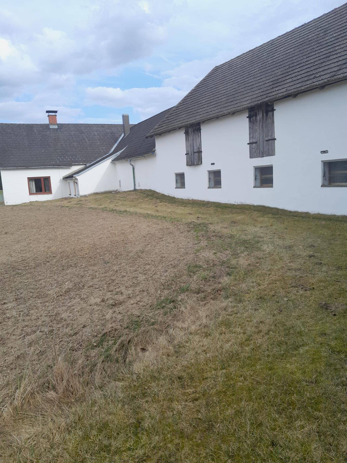 Seitenansicht eines weißen Bauernhauses mit braunen Fensterläden und einem grasbewachsenen Hang im Vordergrund.