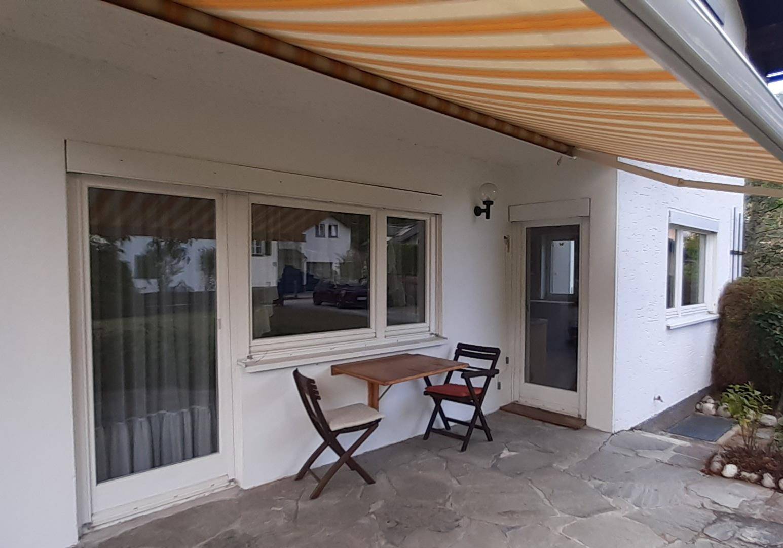 Kleine gepflasterte Terrasse mit Markise und Sitzgelegenheiten direkt am Haus.