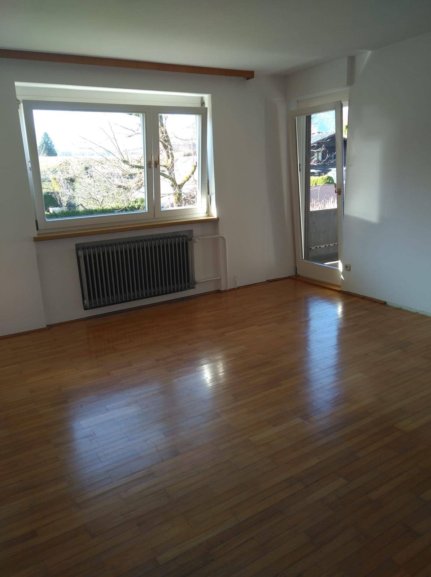 Leeres Zimmer mit Holzboden, großem Fenster und Zugang zu einem kleinen Balkon.