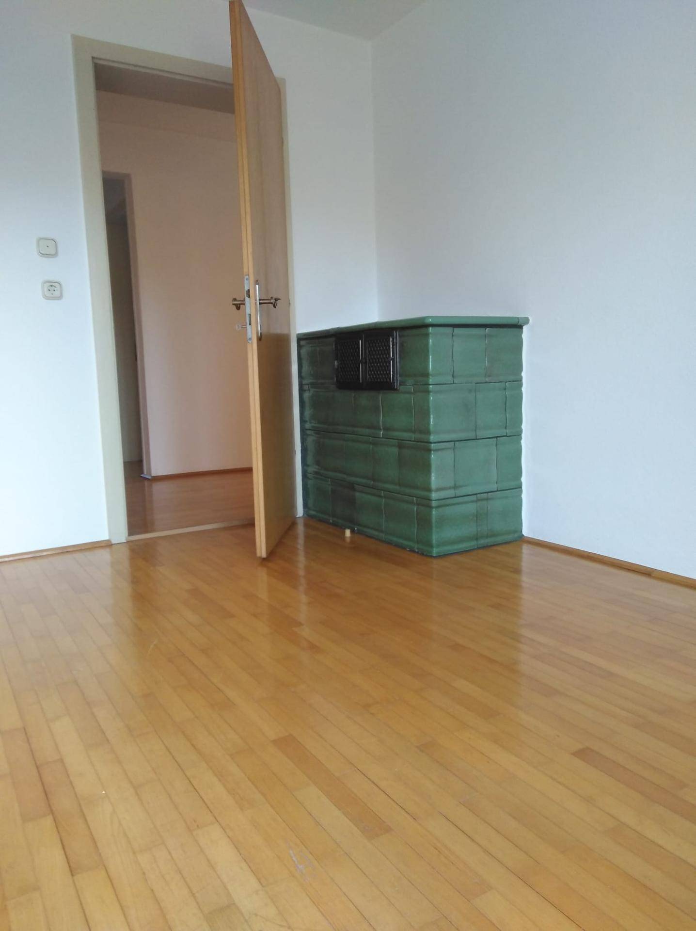 Geräumiges Zimmer mit Holzboden und einem traditionellen grünen Kachelofen.