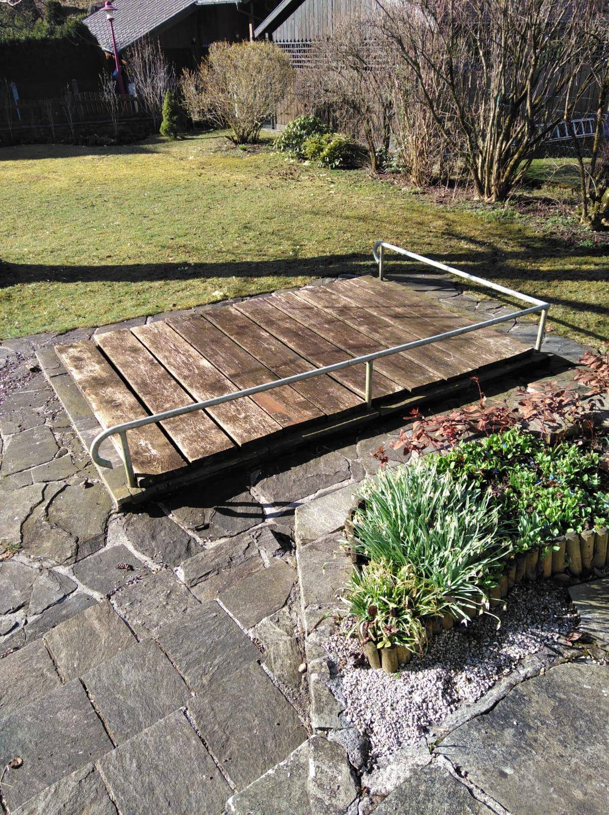 Kleine Holzplattform im Gartenbereich, umgeben von Steinplatten und Pflanzen.