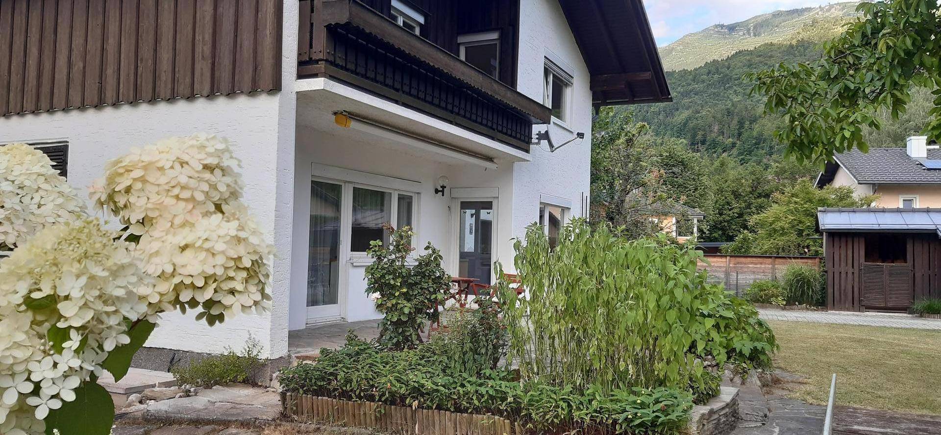 Seitliche Ansicht des Hauses mit Balkon, Terrasse und blühenden Pflanzen im Garten.