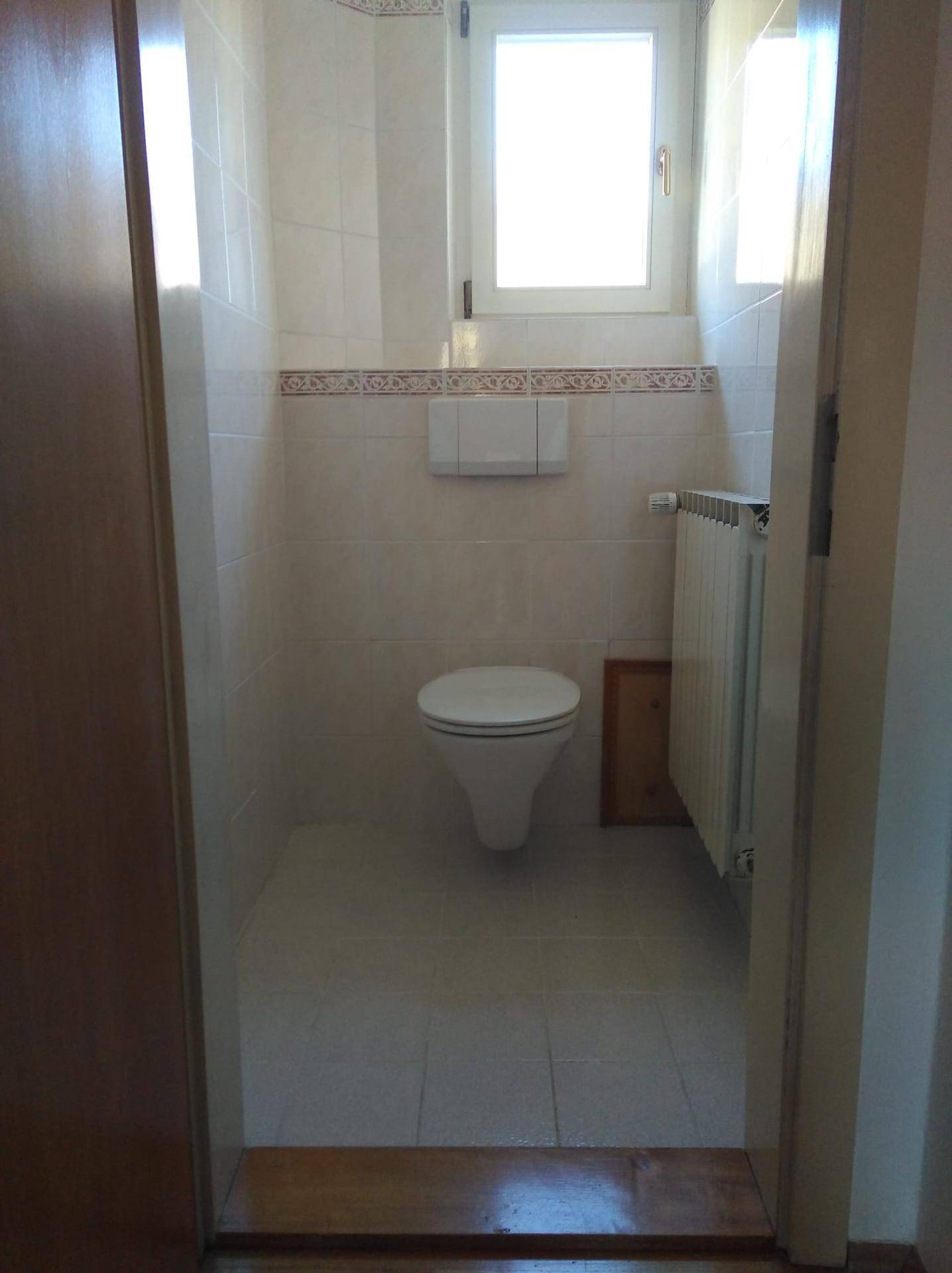 Separates WC mit Wand-WC, Fenster und hellen Fliesen an Wänden und Boden.