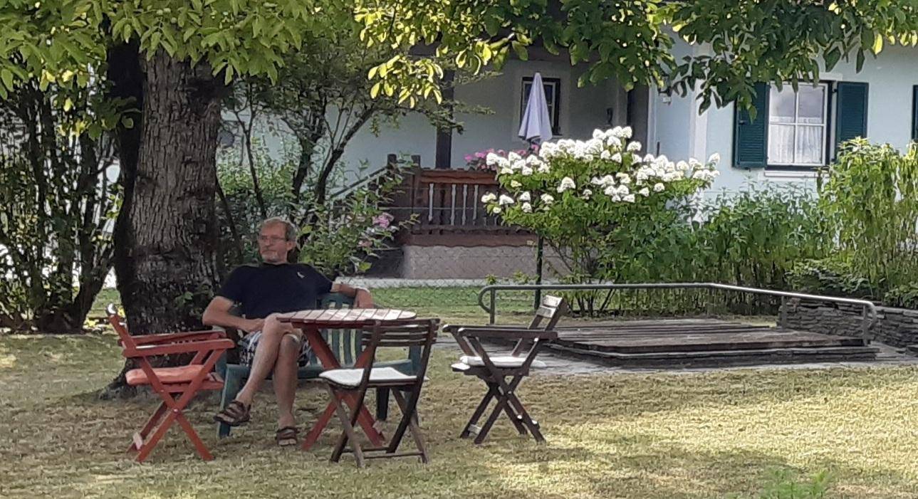 Mann entspannt an einem Gartentisch unter einem Baum mit Blick auf das Haus.