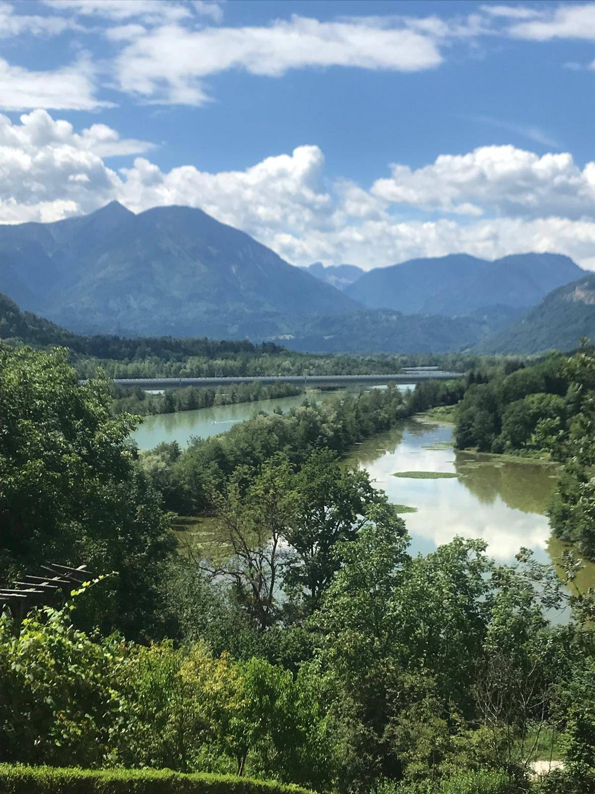 Panoramablick auf einen Fluss, umgeben von üppiger Vegetation und majestätischen Bergen unter blauem Himmel.