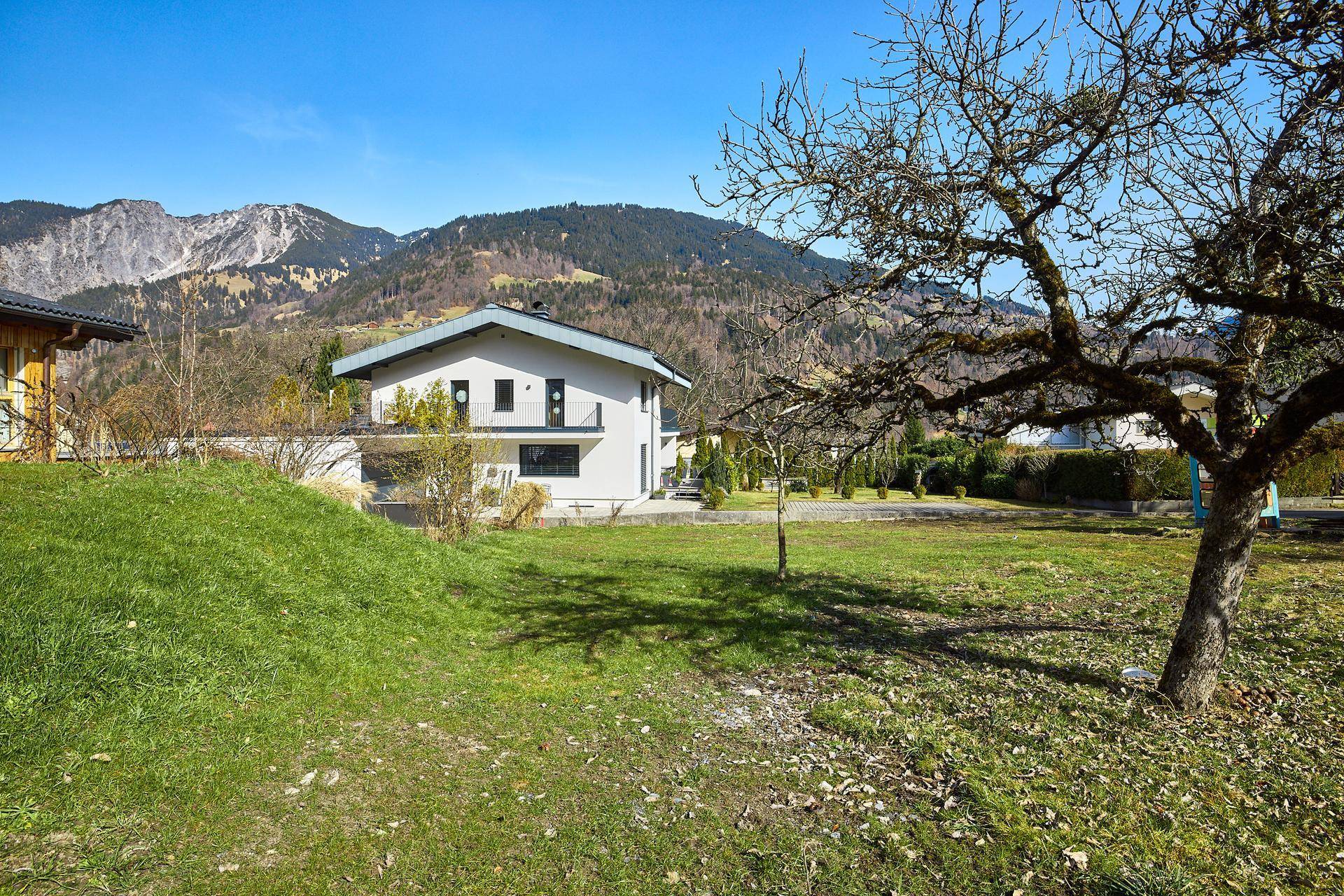 Eine sonnige Wiese mit einem Baum und einem modernen Haus im Hintergrund, umgeben von grünen Hügeln und Bergen.