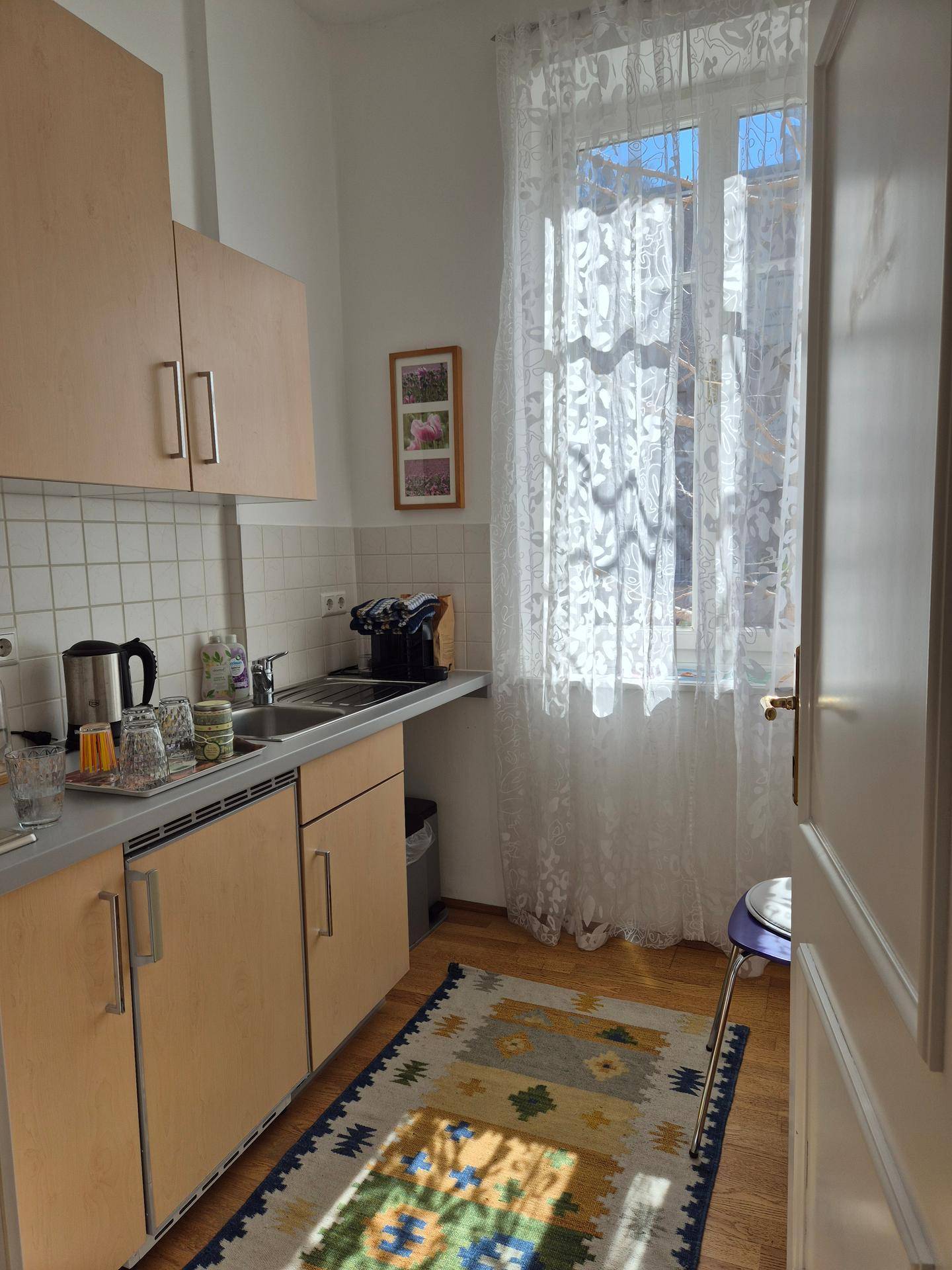 Funktionale Küche mit hellen Holzschränken, Spüle, Arbeitsplatte und einem Fenster mit Spitzenvorhängen.