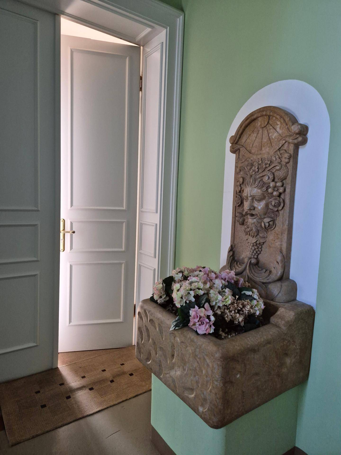 Heller Flur mit einer dekorativen Wandfontäne, Blumen und einer geöffneten weißen Tür, die Licht hereinlässt.