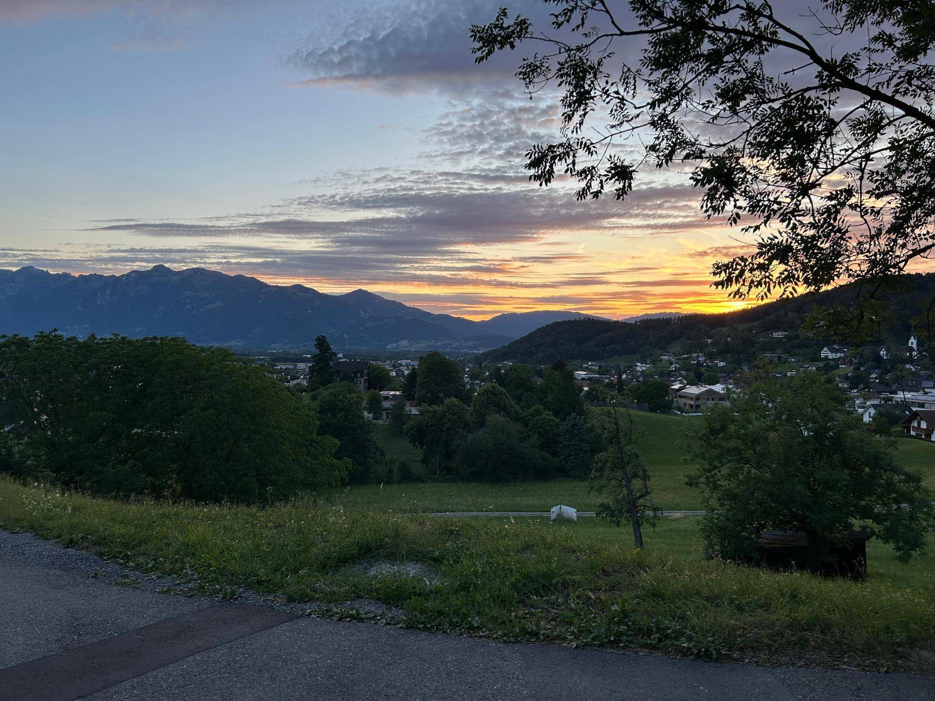 Malerischer Sonnenuntergang über einer Berglandschaft mit grünen Hügeln und einem Dorf im Tal.