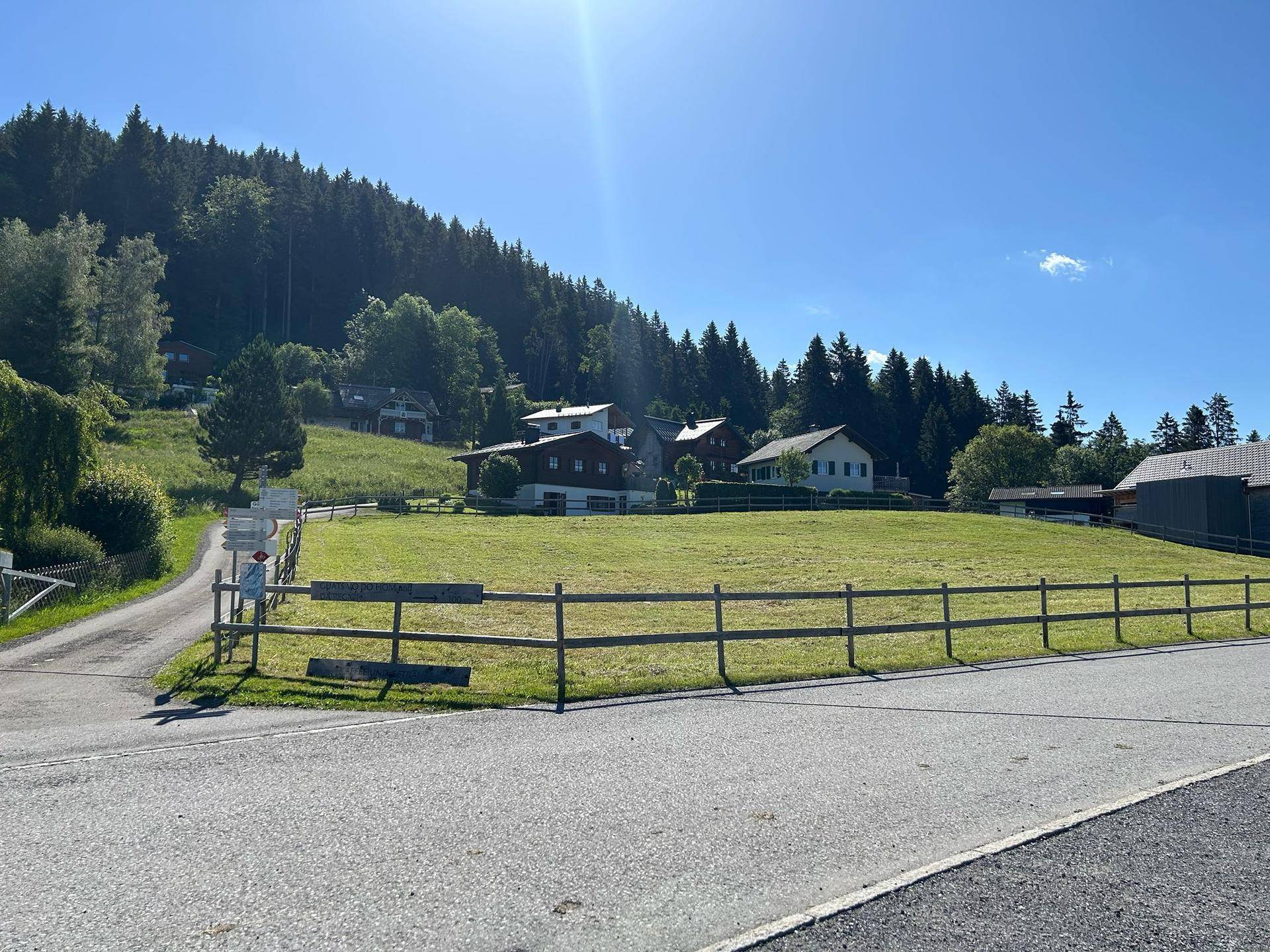 Ländliche Szene mit grüner Wiese, Holzzaun, Straße und Häusern auf einem sonnigen Hügel.