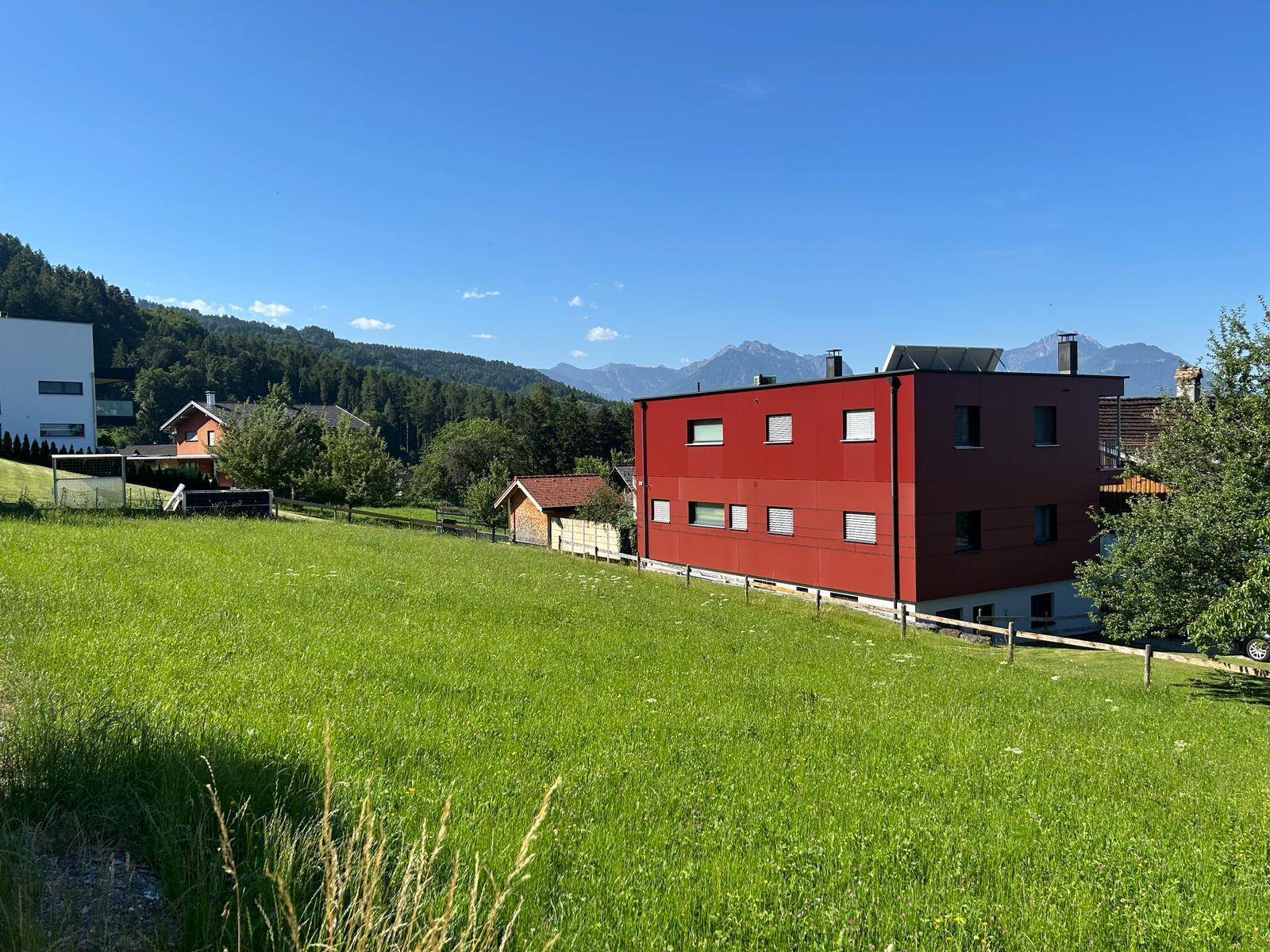 Grüne Wiese mit einem modernen roten Gebäude und majestätischem Bergblick an einem sonnigen Tag.