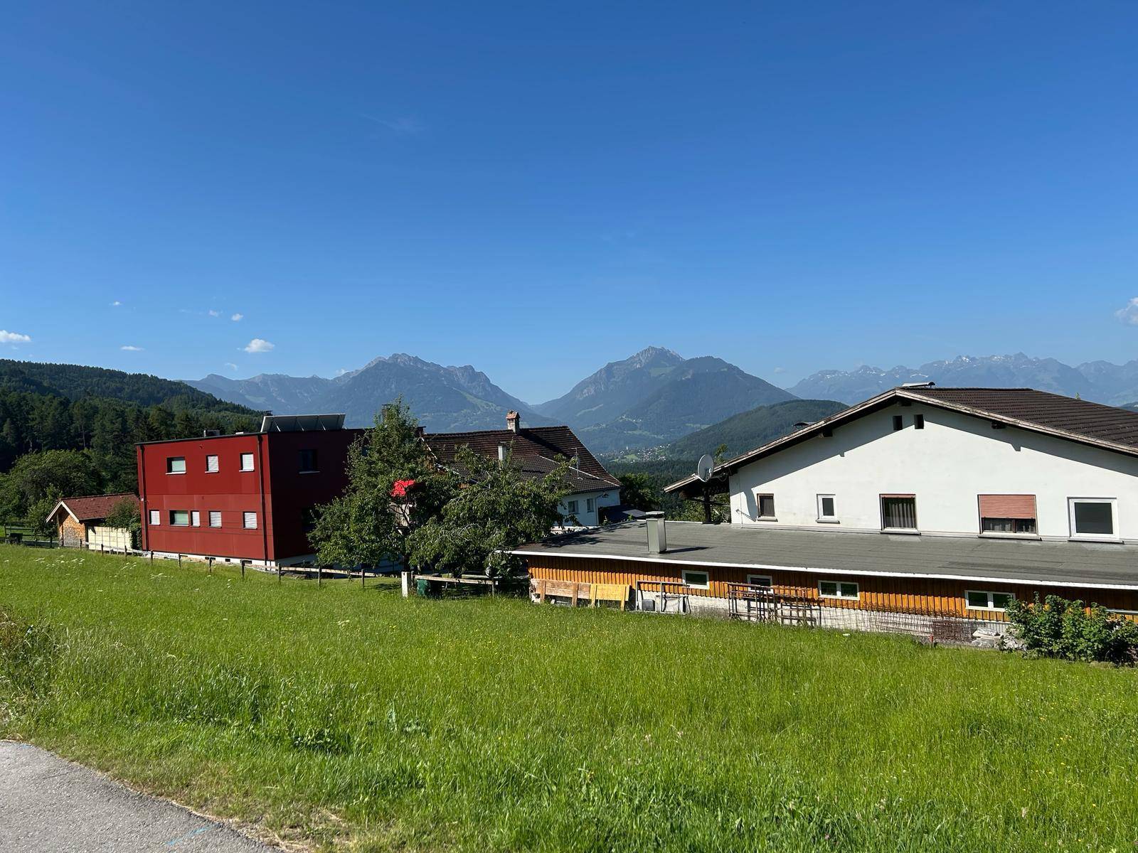 Panoramablick über eine grüne Wiese mit verschiedenen Häusern und einer weiten Berglandschaft im Hintergrund.