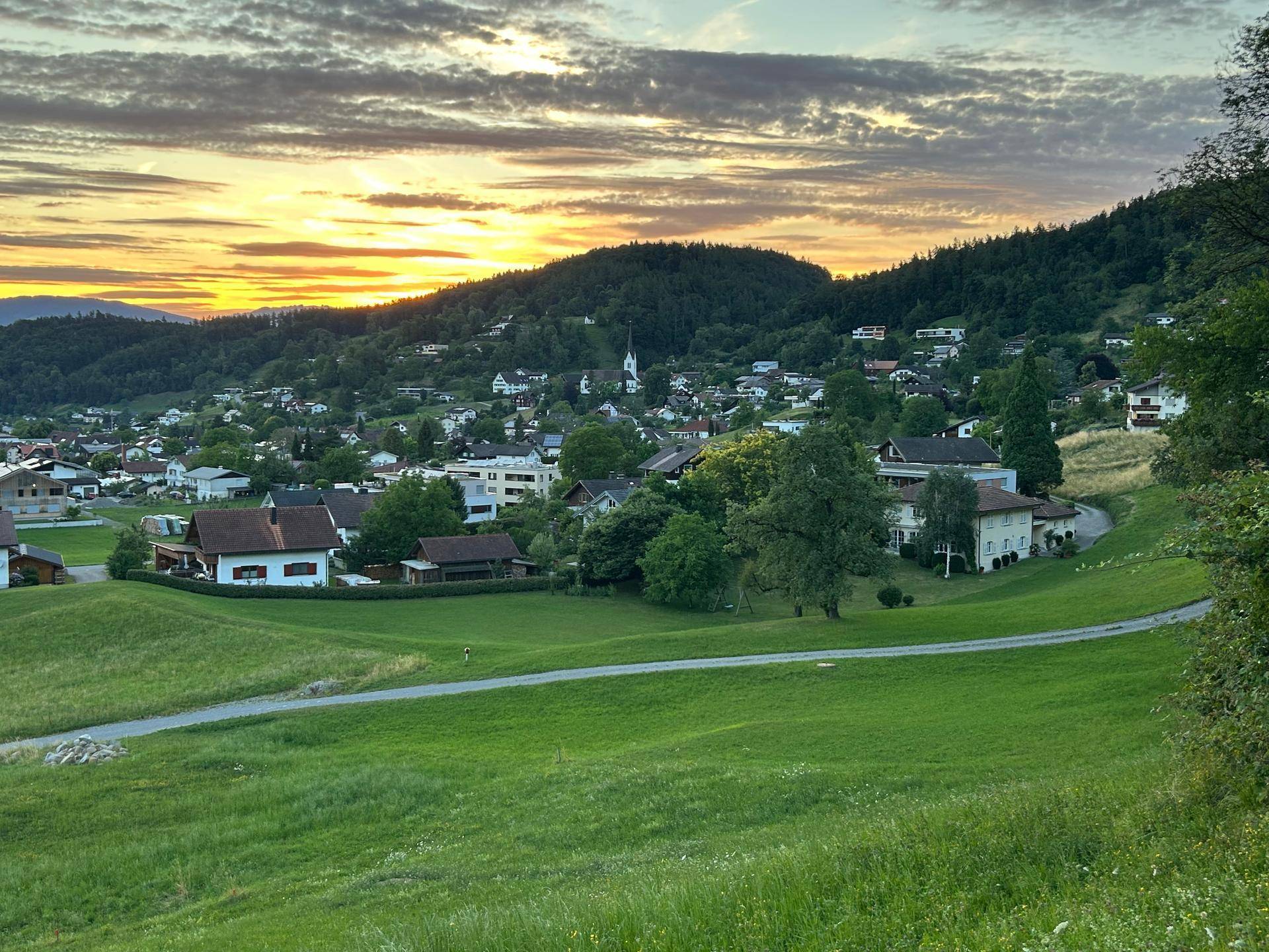 Panoramablick auf ein charmantes Dorf in einem Tal, umgeben von grünen Hügeln bei Sonnenuntergang.
