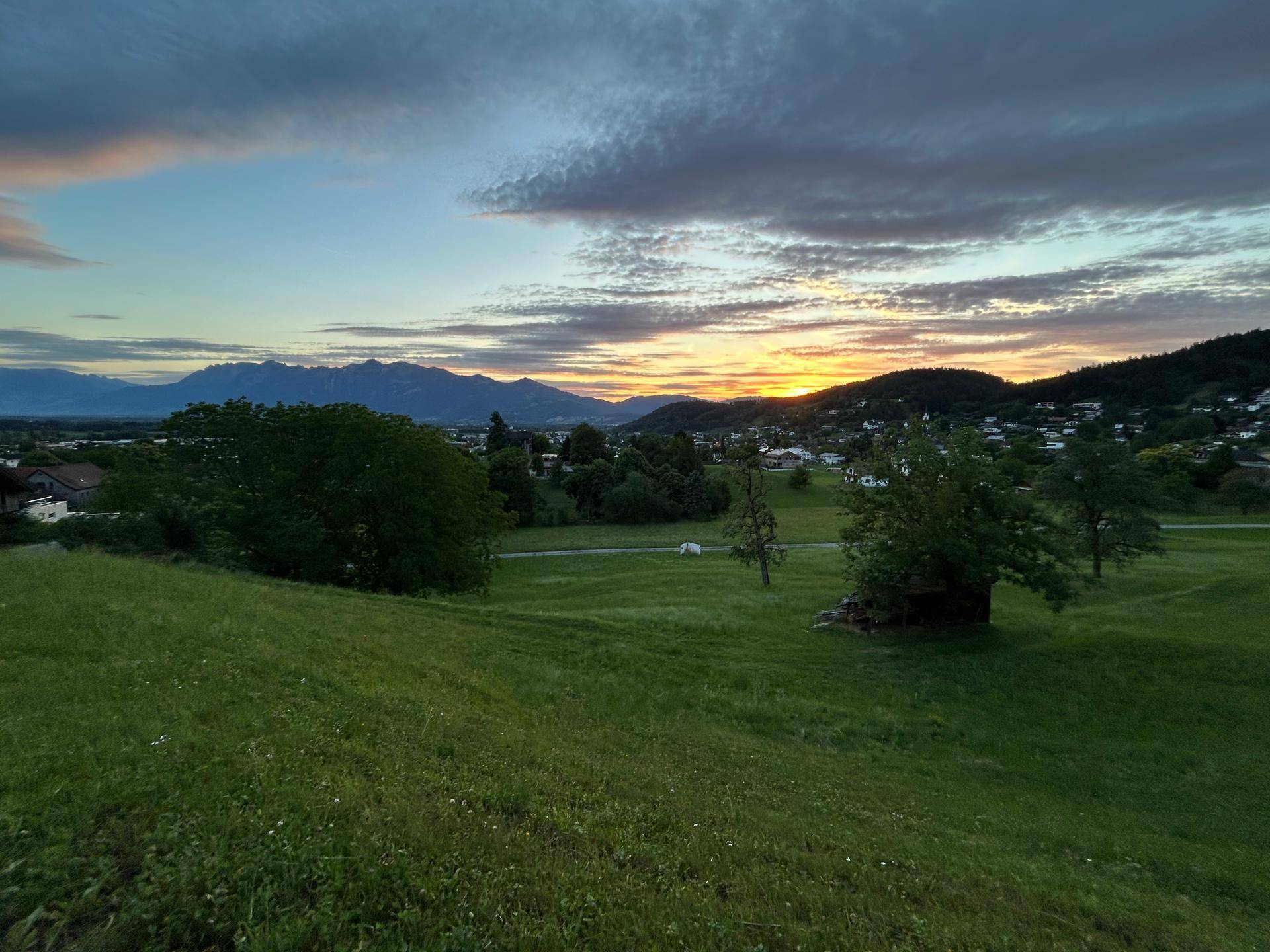 Sonnenuntergang über einer ländlichen Landschaft mit Bergen im Hintergrund und vereinzelten Häusern.