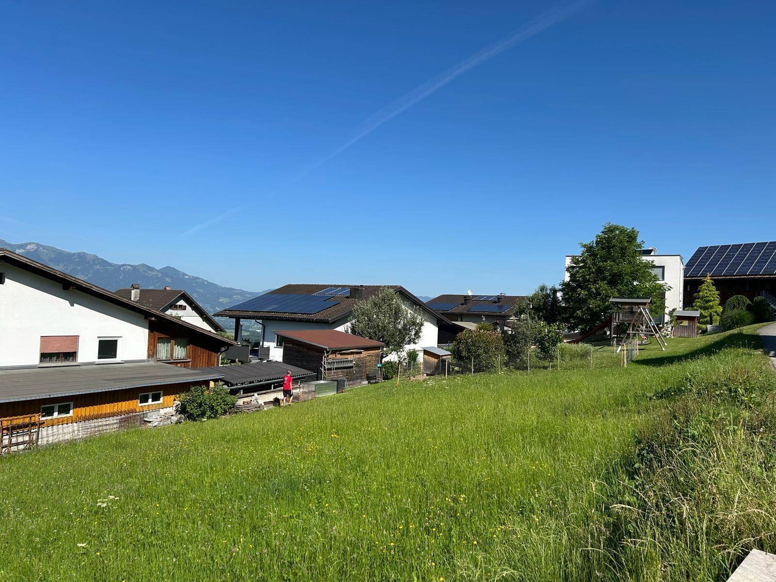 Sonnige Wiese mit Wohnhäusern, Solaranlagen auf Dächern und einem Spielplatz im Hintergrund.