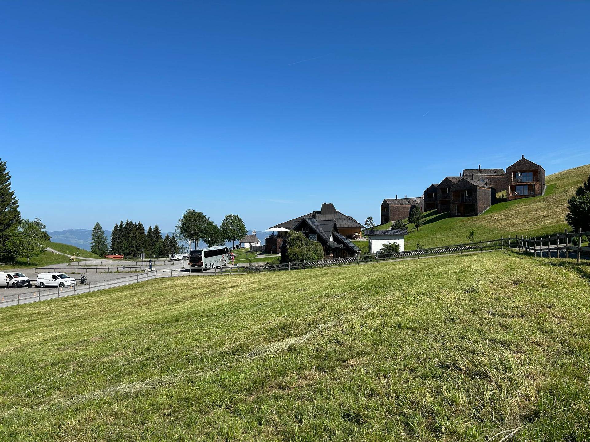 Weite grüne Wiese mit modernen Gebäuden und Parkplätzen im Hintergrund unter blauem Himmel.