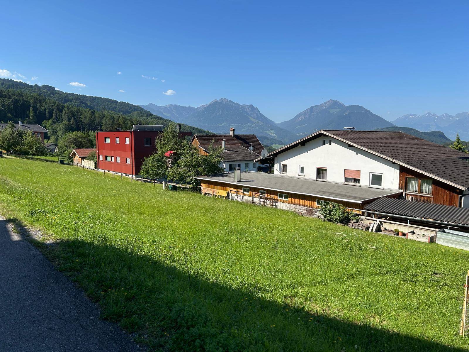 Idyllische grüne Landschaft mit Wohnhäusern und einer malerischen Bergkulisse unter blauem Himmel.