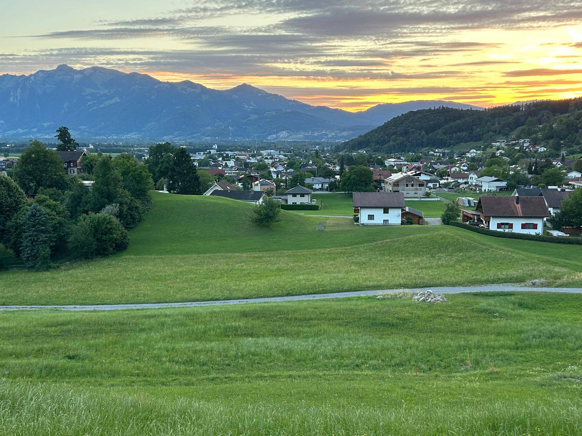Weitläufige Landschaft mit verstreuten Häusern, grünen Feldern und einer beeindruckenden Bergkulisse bei Sonnenuntergang.