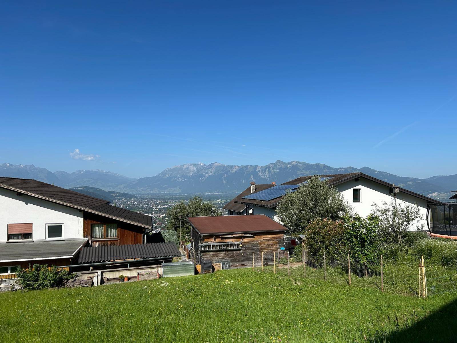 Weitblick über eine grüne Wiese auf Häuser, eine Stadt im Tal und eine beeindruckende Bergkette am Horizont.