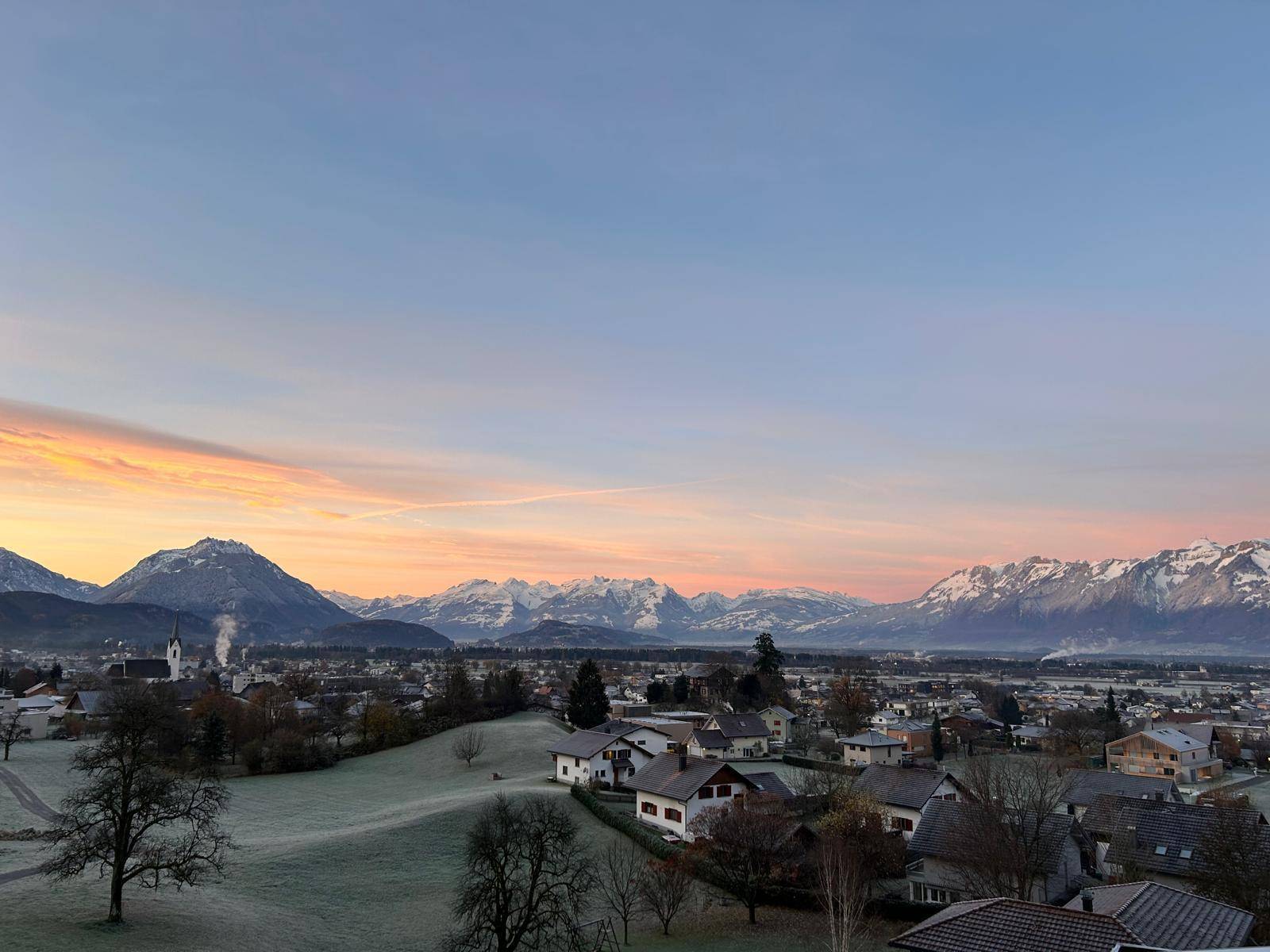 Atemberaubender Sonnenuntergang über einem Dorf mit schneebedeckten Bergen und warmen Himmelsfarben.