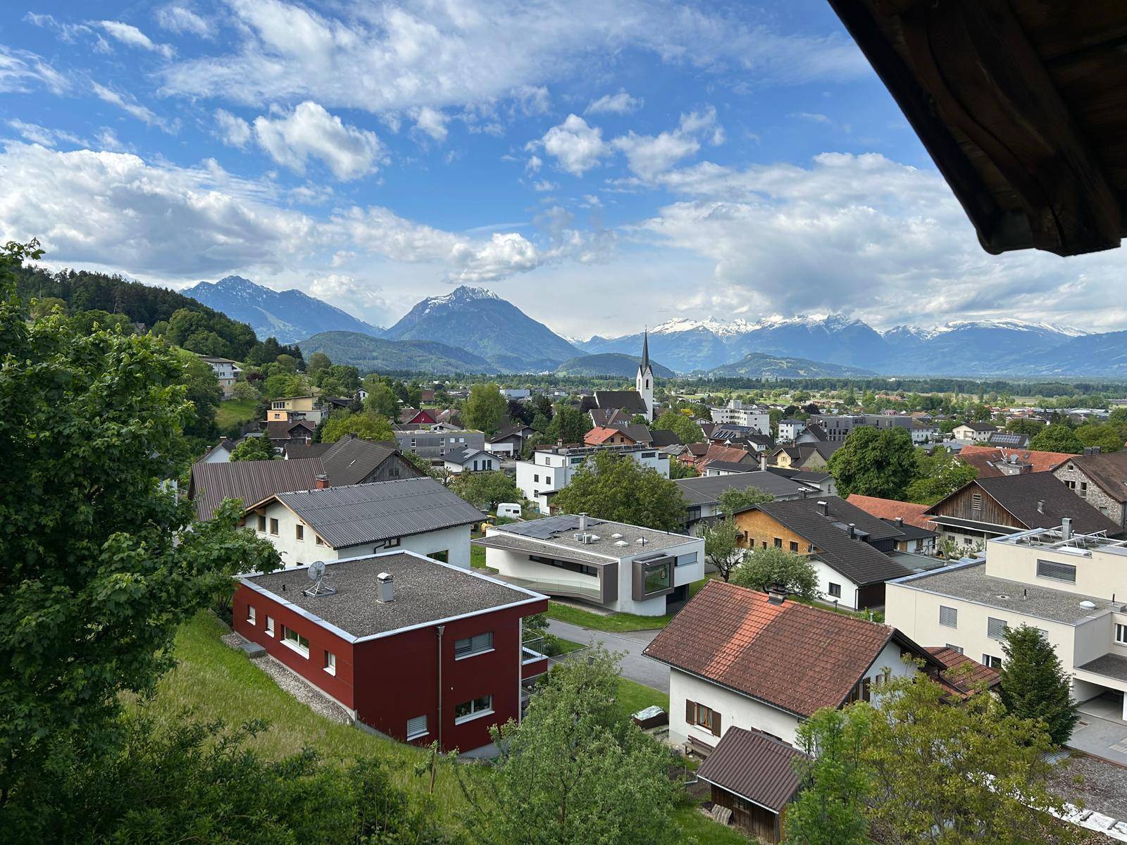 Blick über ein dicht besiedeltes Dorf mit verschiedenen Häusern und einer Kirche vor alpiner Kulisse.