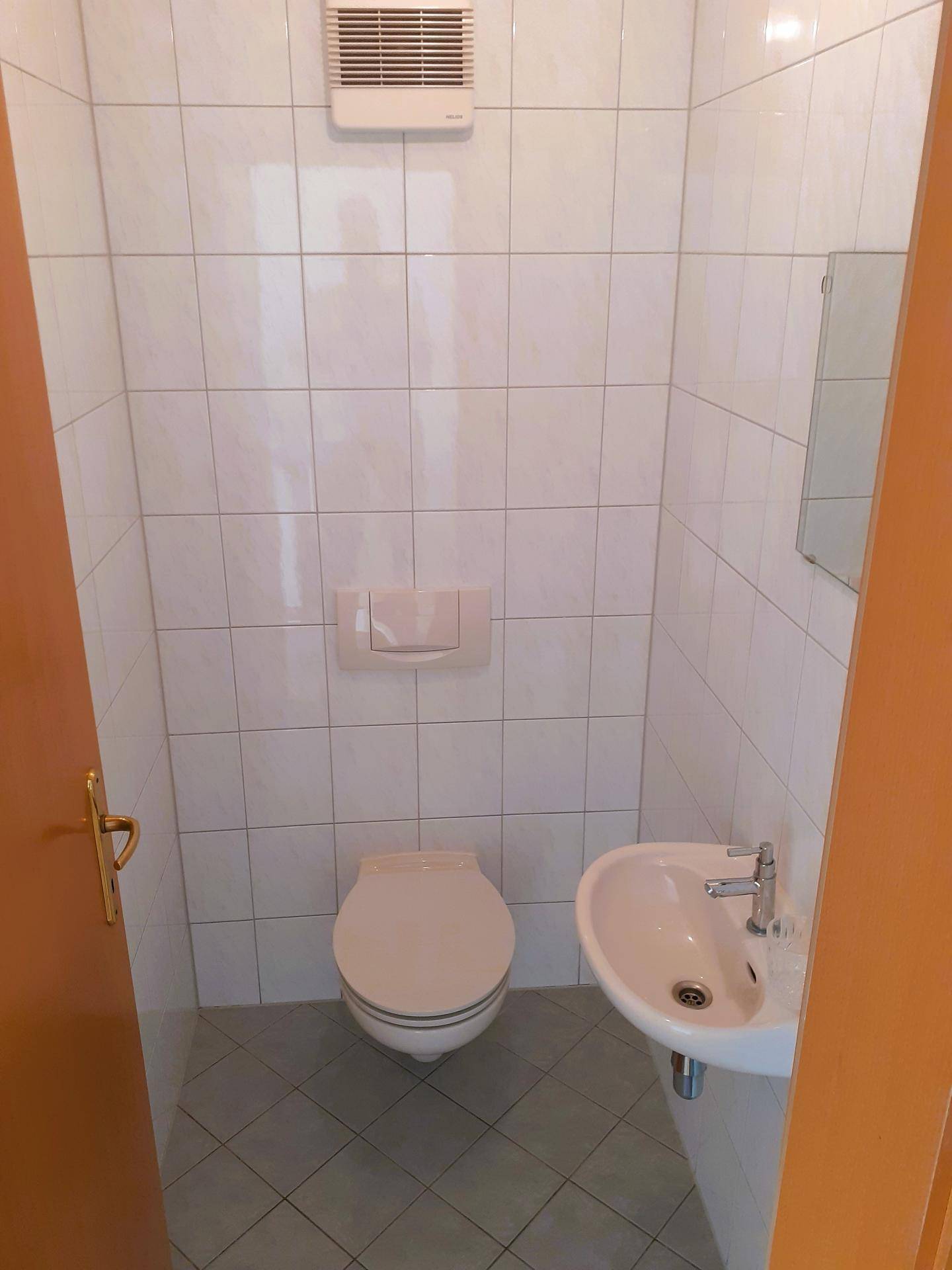 Gäste-WC mit Wand-WC, kleinem Waschbecken und weißen Fliesen, ausgestattet mit Abluftventilator.
