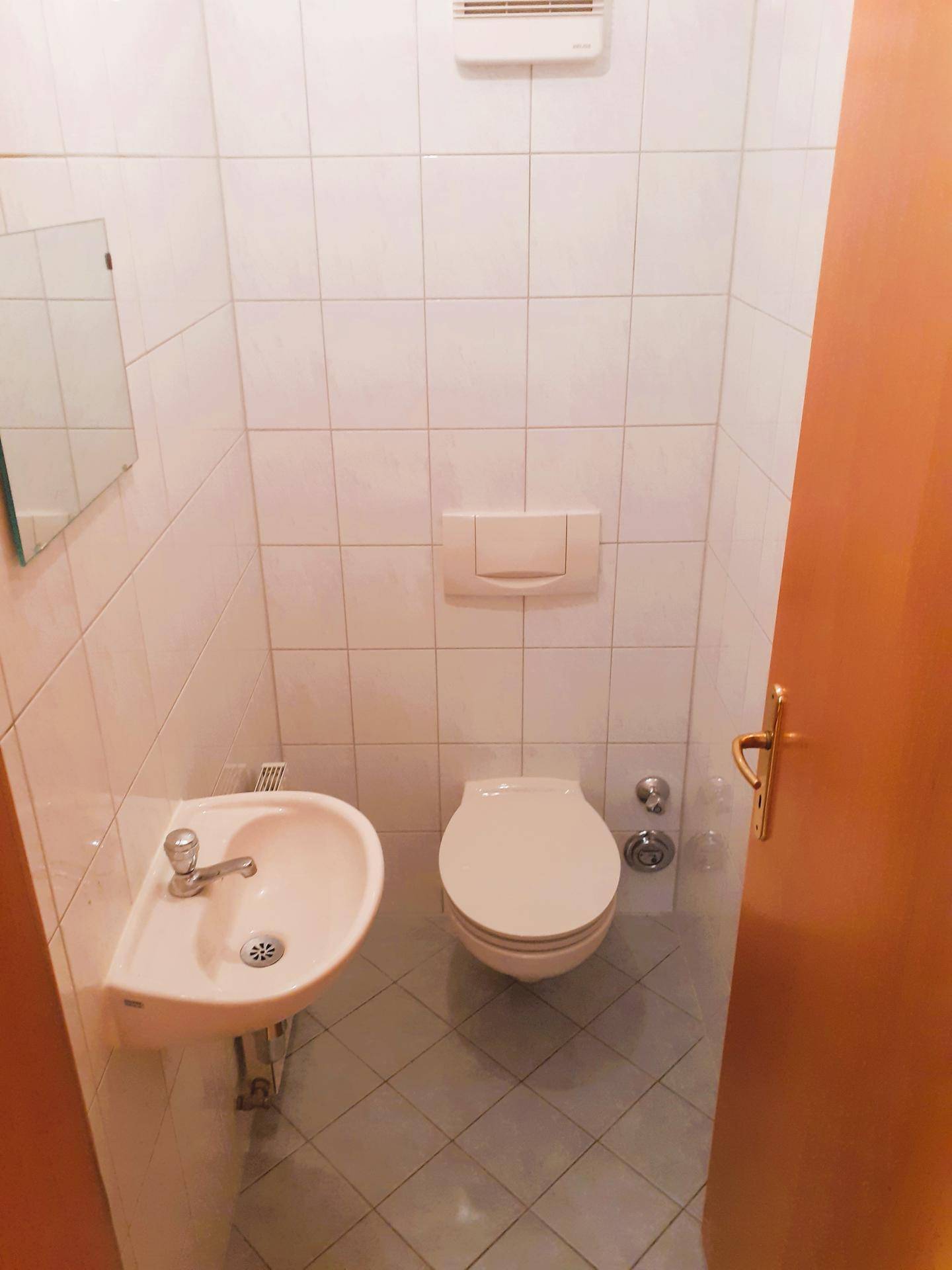 Gäste-WC mit Wand-WC, kleinem Waschbecken und weißen Fliesen, ausgestattet mit Abluftventilator.