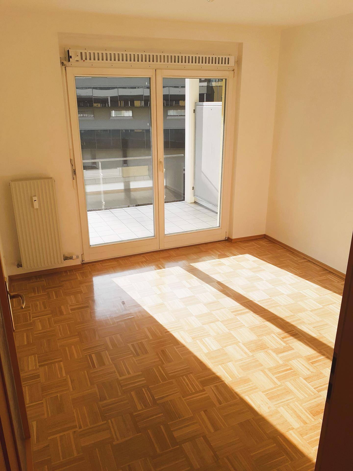 Helles Zimmer mit Parkettboden und großen Fenstern, die den Raum mit Sonnenlicht füllen.