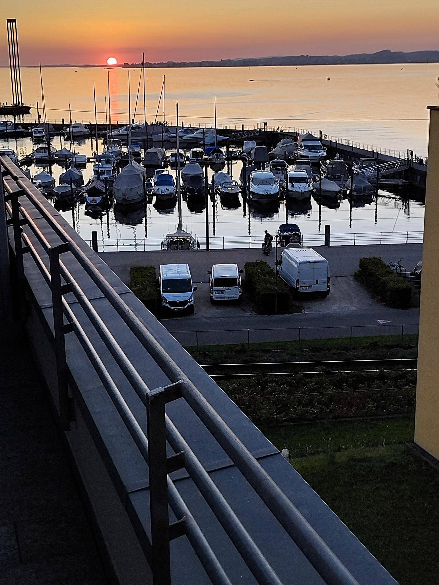 Blick vom Balkon auf einen Yachthafen bei Sonnenuntergang mit ruhigem Wasser und parkenden Fahrzeugen.