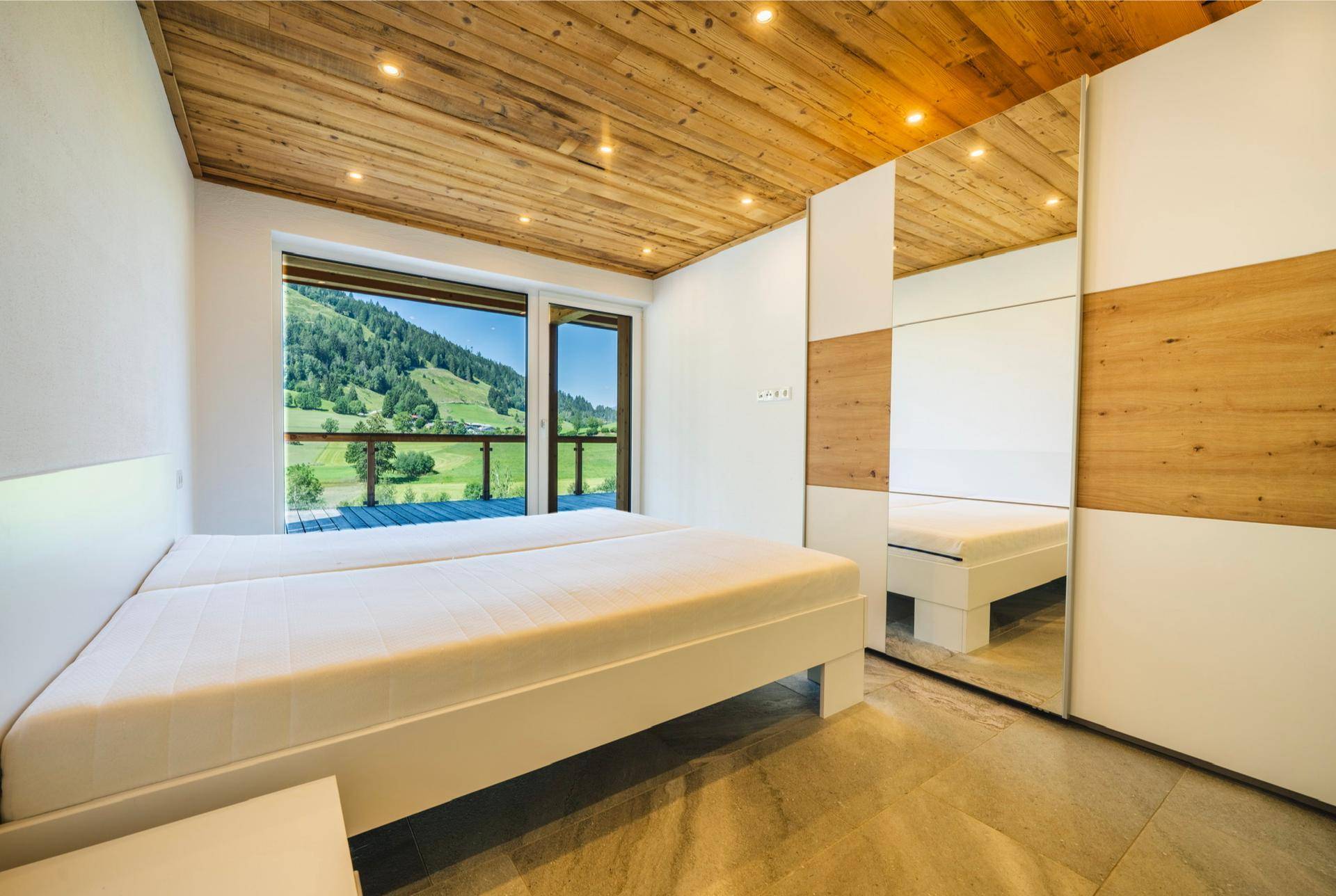 Schlafzimmer mit Holzbalkendecke, großem Fenster und Zugang zu einem Balkon mit Bergblick.