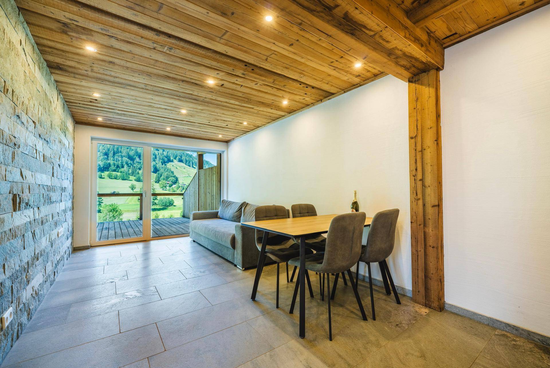 Gemütlicher Wohn- und Essbereich mit Holzbalkendecke, Steinwand und Zugang zu einem Balkon.